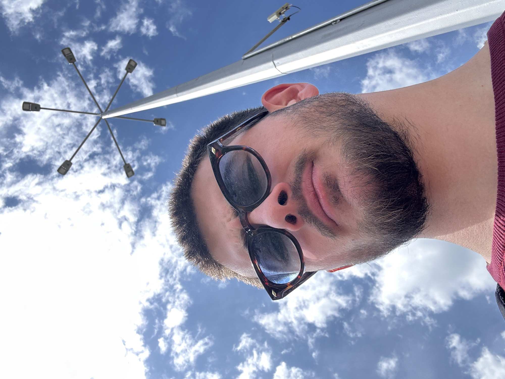The image depicts a low-angle upward view of a man in sunglasses under a blue, cloudy sky. A light pole with multiple arms extending outwards is visible above him, as well as an overhead bar with a possible security camera attached to it. The location appears to be somewhere in the city of Sofia, Bulgaria.