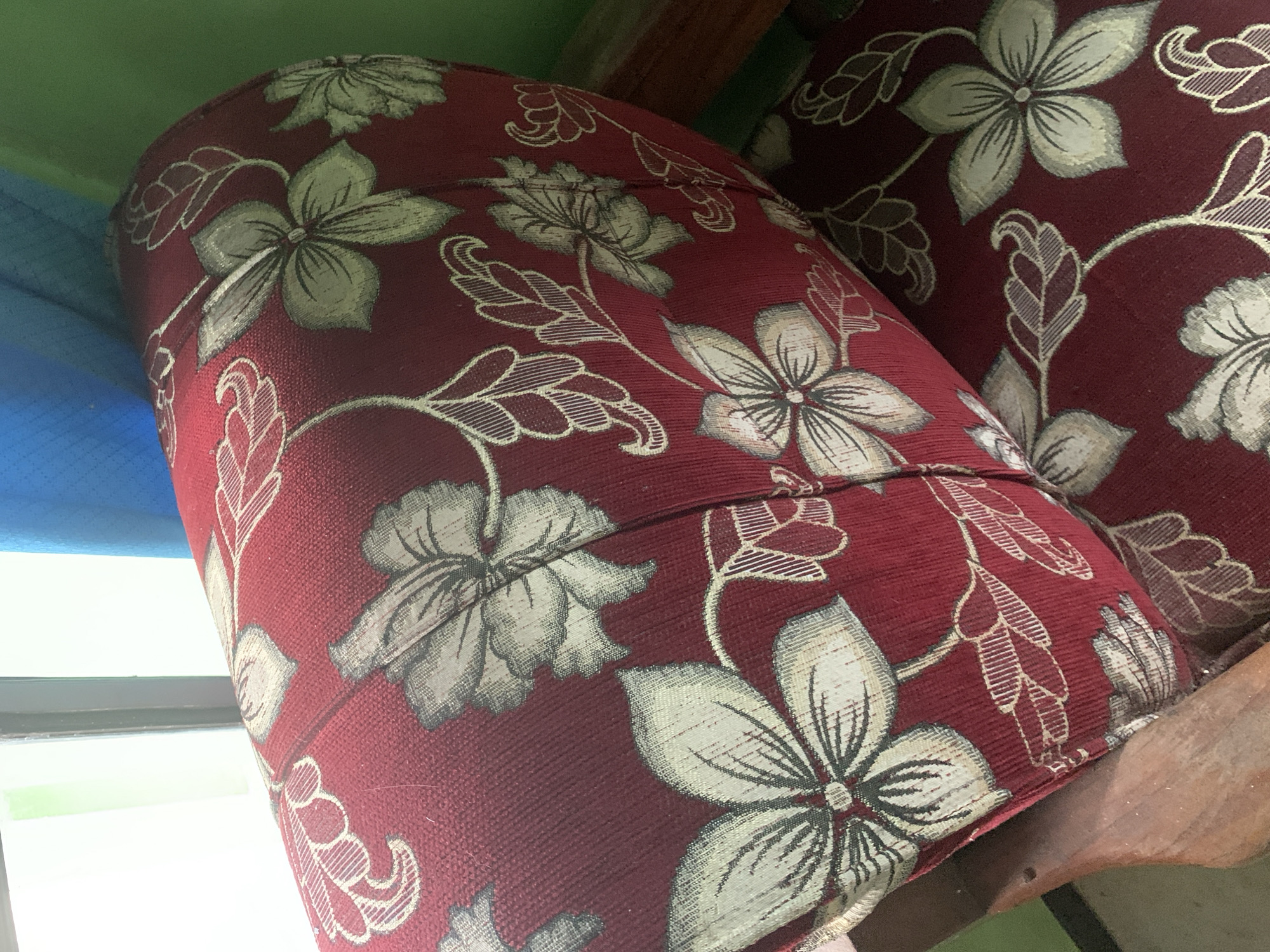 The image shows a close-up of a cushioned sofa with a red fabric and a floral pattern of light green and cream flowers and leaves. The sofa appears to be made of wood, and the cushioning is a thick, textured material. In the background, a window with light filtering through and a blue curtain are visible. The environment suggests an indoor setting, possibly a living room. The specific location in Dukuh Mojo, Indonesia, cannot be determined from the image alone, as there are no identifying landmarks or signs present. The scene is static, with no people or actions depicted.