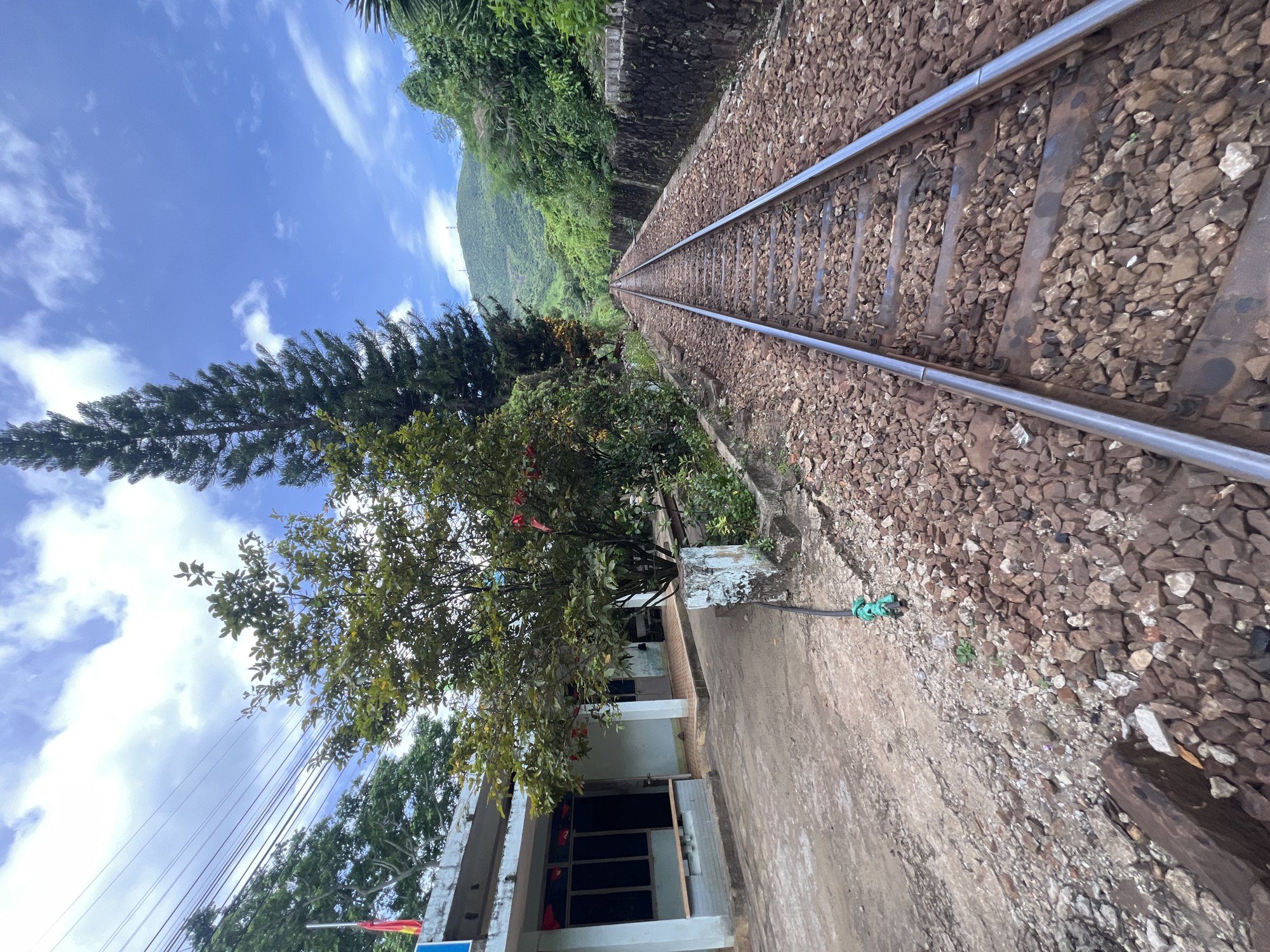 An outdoor, ground-level, eye-level, wide-angle shot captures a railway track stretching into the distance. The track, made of steel rails and wooden sleepers, is flanked by a dense arrangement of brown and grey gravel. To the left of the track, a low-rise, light blue building with a white roof features several windows and doors. Adjacent to the building and between it and the railway track is a small tree with green leaves and scattered red decorations. A tall, thin evergreen tree stands behind the small tree. A stone wall runs alongside the railway track on the right. The background consists of lush green hills and a bright blue sky with scattered white clouds. The scene suggests a rural or suburban setting, likely on the outskirts of Huế, Vietnam, given the architectural style of the building and the vegetation. There are no people or animals visible in the image. The overall impression is one of a quiet, somewhat dated, transportation hub in a verdant landscape.