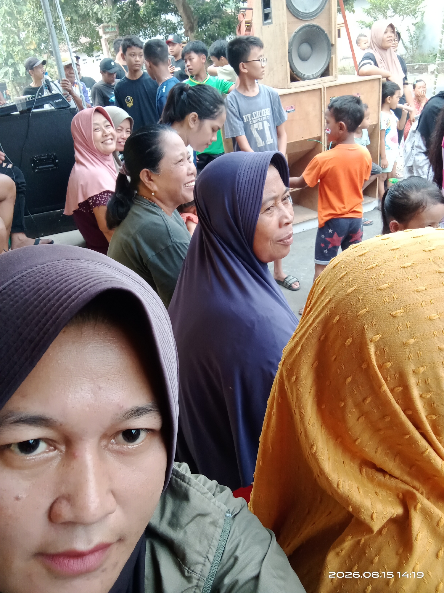 A group of people are gathered outdoors in what appears to be a casual event, possibly a community gathering or outdoor performance, in Banyuarang, Indonesia. The scene is dominated by large speaker boxes, suggesting a sound system is set up. In the foreground, a person wearing a dark hijab is taking a selfie, looking directly at the camera. To their right, another person in a purple hijab is facing left, with their mouth slightly open as if speaking. Partially visible behind them is a person wearing a bright yellow, textured garment.

Further back, several other individuals are visible, including women in headscarves and men and children. One woman in a pink hijab is smiling and looking to her left. A child in an orange shirt with star-patterned shorts is standing near the speaker boxes, holding something small. The environment appears to be outdoors, with trees and foliage visible in the background, indicating a possibly rural or semi-rural setting within Banyuarang. The lighting suggests it is daytime. A timestamp on the bottom right of the image reads "2026.08.15 14:19", which is likely a future date and time stamp from the camera.