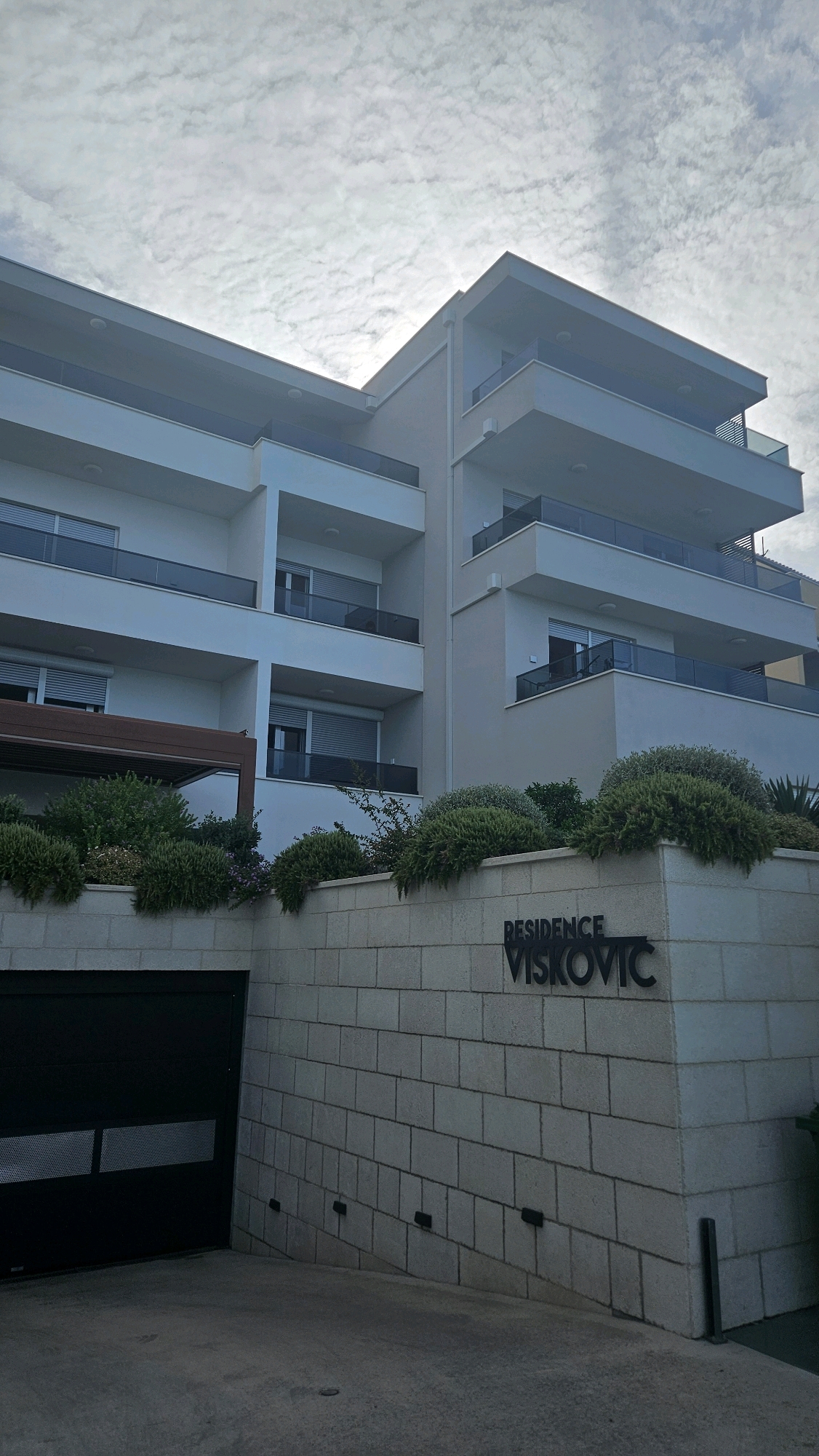 The image shows the exterior of a modern, multi-story apartment building, labeled "RESIDENCE VISKOVIC". The building is light-colored, with multiple balconies featuring glass railings. In the foreground, there is a stone wall with the building's name prominently displayed. To the left of the wall is a large, black garage door. Lush green bushes and some smaller plants are situated on top of the stone wall, partially obscuring the lower floors of the building. The sky above is partly cloudy. The scene appears to be in a residential area, likely a coastal town given the architectural style and vegetation. The location is Općina Tučepi, Croatia.