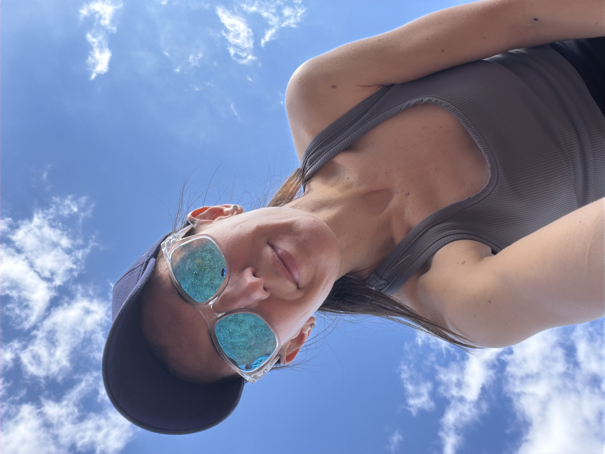 A woman wearing a navy blue baseball cap and clear-framed sunglasses with blue mirrored lenses is looking up at the sky. She has dark hair and is wearing a gray tank top. The background is a bright blue sky with scattered white clouds. The image appears to be taken from a low-angle perspective, looking up at the woman. There is no indication of the city of Chernushka, Russia, in the image.