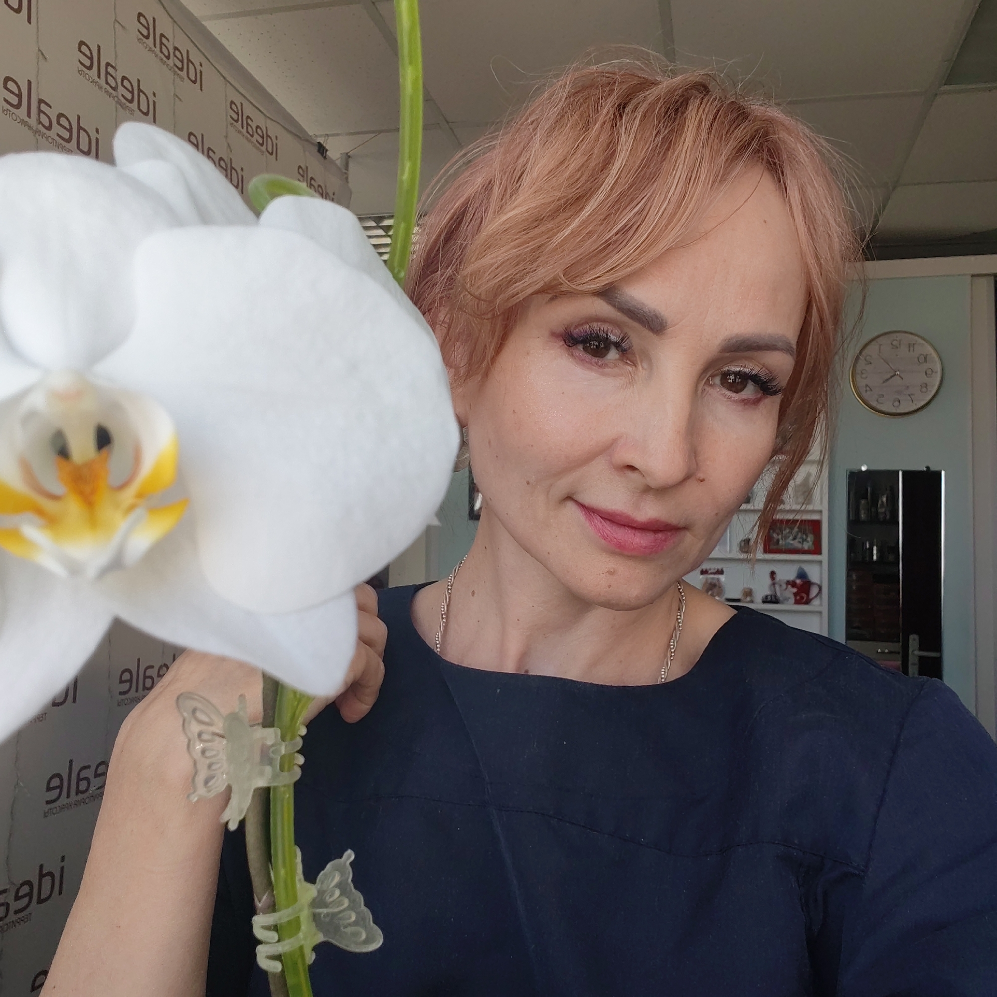 The image is a medium close-up selfie of a fair-skinned woman with short, reddish-blonde hair. She's positioned slightly to the right of the frame, holding a large white orchid flower with a yellow center on the left side of the frame. The flower is partially obscuring her left shoulder and arm, and the stem is secured with two clear plastic butterfly clips.
She is wearing a dark blue or black top with a simple, boat-neck design. A delicate silver necklace is visible around her neck. Her makeup includes defined eyebrows, dark mascara, and rosy pink lipstick.
In the background, there is a wall with repeating text that appears to be a brand name or logo. Behind the woman and slightly to her right, there is a glimpse of an interior room, including a light blue wall on which is a gold-rimmed clock. Also visible in the background is shelving that contains various small items. The time on the clock is 10:55.
The location is Chaikovsky, Russia.
