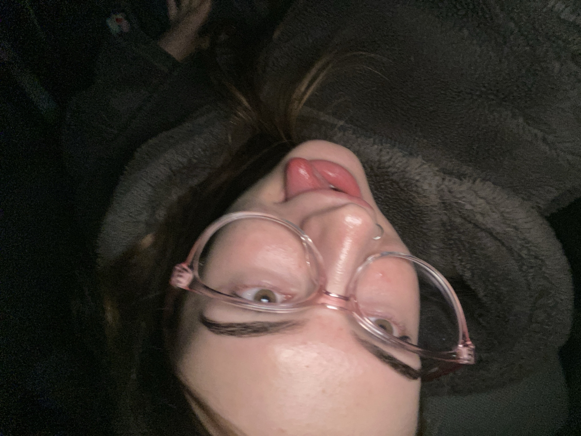 An upside-down close-up selfie captures a person looking directly at the camera with a playful expression. The subject is a light-skinned individual, likely female, with long brown hair visible above their head. They are wearing light pink-framed glasses, which are prominent in the foreground, and have light-colored eyes (appearing hazel or light brown). Their eyebrows are dark and well-defined. A small silver hoop nose piercing is visible on their right nostril.
The person's tongue is sticking out and curved upwards, contributing to a humorous and silly demeanor. Their face shows some freckles. They appear to be wearing or covered by a dark, fuzzy, fleece-like material, possibly a blanket, hoodie, or robe, which fills the background. The lighting is dim, suggesting an indoor setting or low light conditions, with shadows creating a dark and soft background. No specific location cues are discernible beyond an indoor environment. There is no visible text in the image.