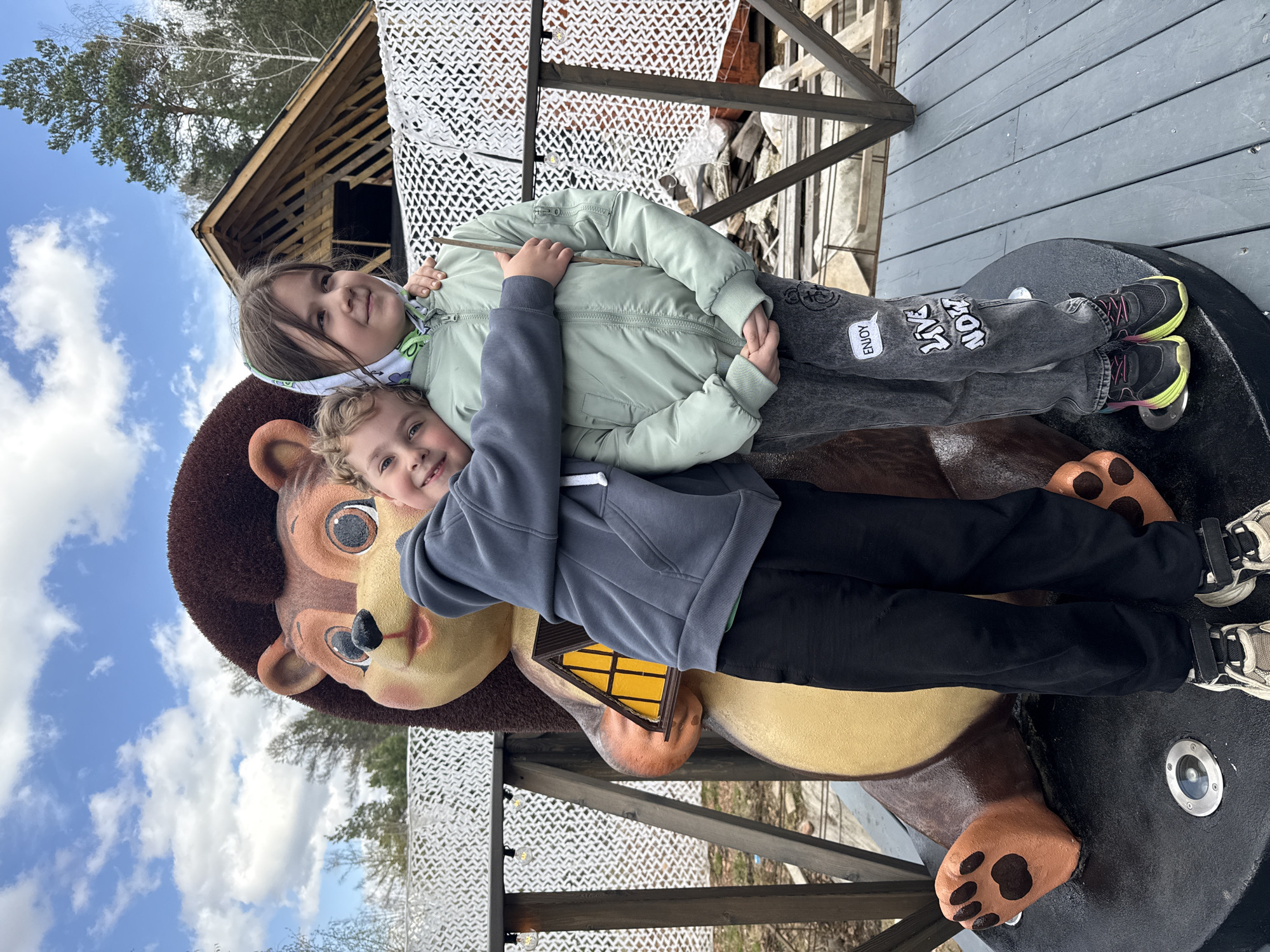 Two children, a boy and a girl, are positioned on a brown bear statue. The boy is lying on the bear's head, facing forward, while the girl is on the bear's back, facing the opposite direction. The statue has paw prints and what appears to be a small yellow house. The backdrop includes a wooden building under construction and some trees, with a blue sky and scattered clouds visible above. The photograph was taken in Divnogorsk, Russia.