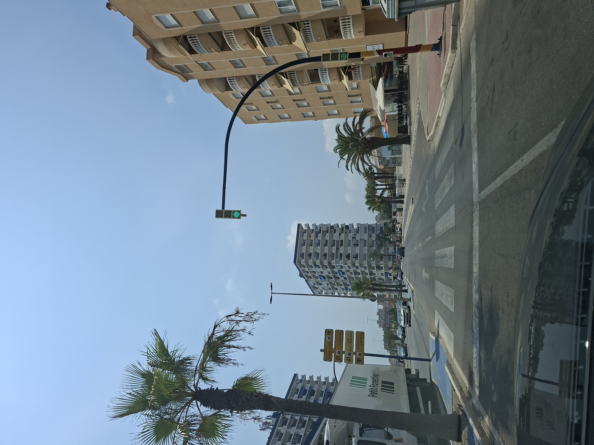 The image shows a street scene in La Manga Del Mar Menor, Spain, on a clear day with a blue sky. A wide, paved road with multiple lanes and a pedestrian crossing is visible. Traffic lights are present, with a green light illuminated on the main signal. Palm trees line the street, some of which are mature and tall. Modern, multi-story apartment buildings with balconies are prominent on both sides of the road. On the left side, a white truck with green lettering that reads "Petit Forestier" is parked. Yellow directional signs indicating hotels such as "Hotel Benidorm La Manga," "Hotel Doblemar," "Hotel Bahia La Manga" and "Aparthotel Londres" are also visible. Cars are driving on the road. On the right side of the road, there are outdoor seating areas for a cafe or restaurant with umbrellas providing shade. The overall environment suggests a coastal resort town.