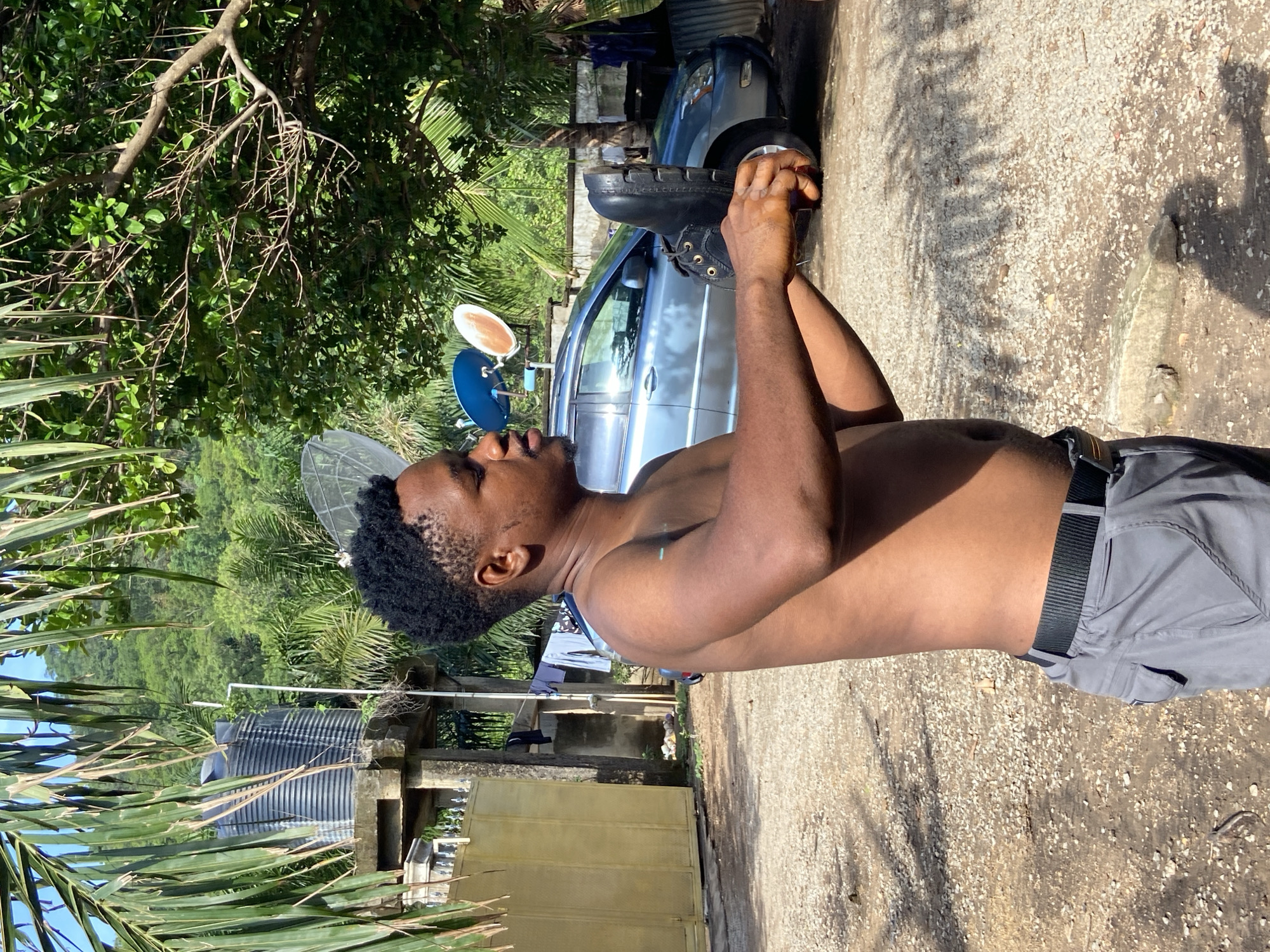 The image shows a man outdoors in Mararaba, Nigeria. He is shirtless, wearing grey shorts with a black belt. He is holding a black leather boot. A light blue car is in the background. A water tank and a satellite dish are also visible. The environment includes trees and foliage.