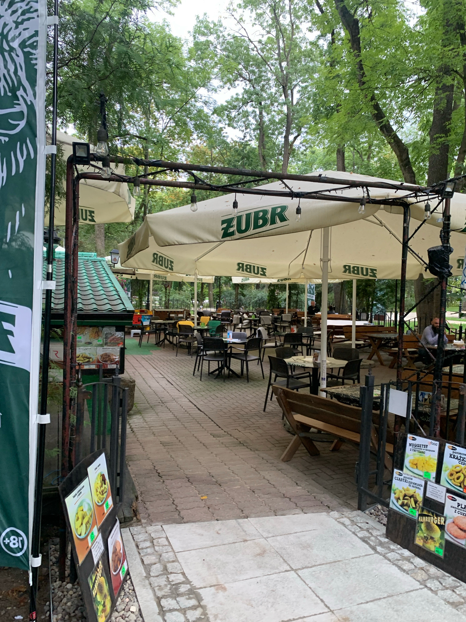 The image depicts an outdoor seating area of a restaurant or cafe, likely located in the city of Białystok, Poland, based on the presence of the ZUBR brand (a popular Polish beer). The scene is set amidst lush green trees, suggesting a park or a shaded courtyard.
Dominating the foreground and midground are several large, cream-colored umbrellas bearing the "ZUBR" logo, providing ample shade. Underneath these umbrellas, numerous tables and chairs are arranged on a cobblestone and brick pathway. Some tables are set with placemats. Wooden benches are also visible, offering additional seating.
To the left, a tall green banner with a white graphic (possibly a logo or advertisement) is partially visible, along with a sign displaying food items in a carousel format. Next to this, a small food stand with a green, tiled roof is present, displaying various dishes behind glass.
In the background, more trees create a natural canopy, and a few individuals are seated at tables, engaging in dining or conversation. The overall atmosphere appears relaxed and casual, characteristic of an outdoor dining establishment in a pleasant setting. The lighting suggests it is daytime, possibly overcast or late afternoon, given the diffused light.