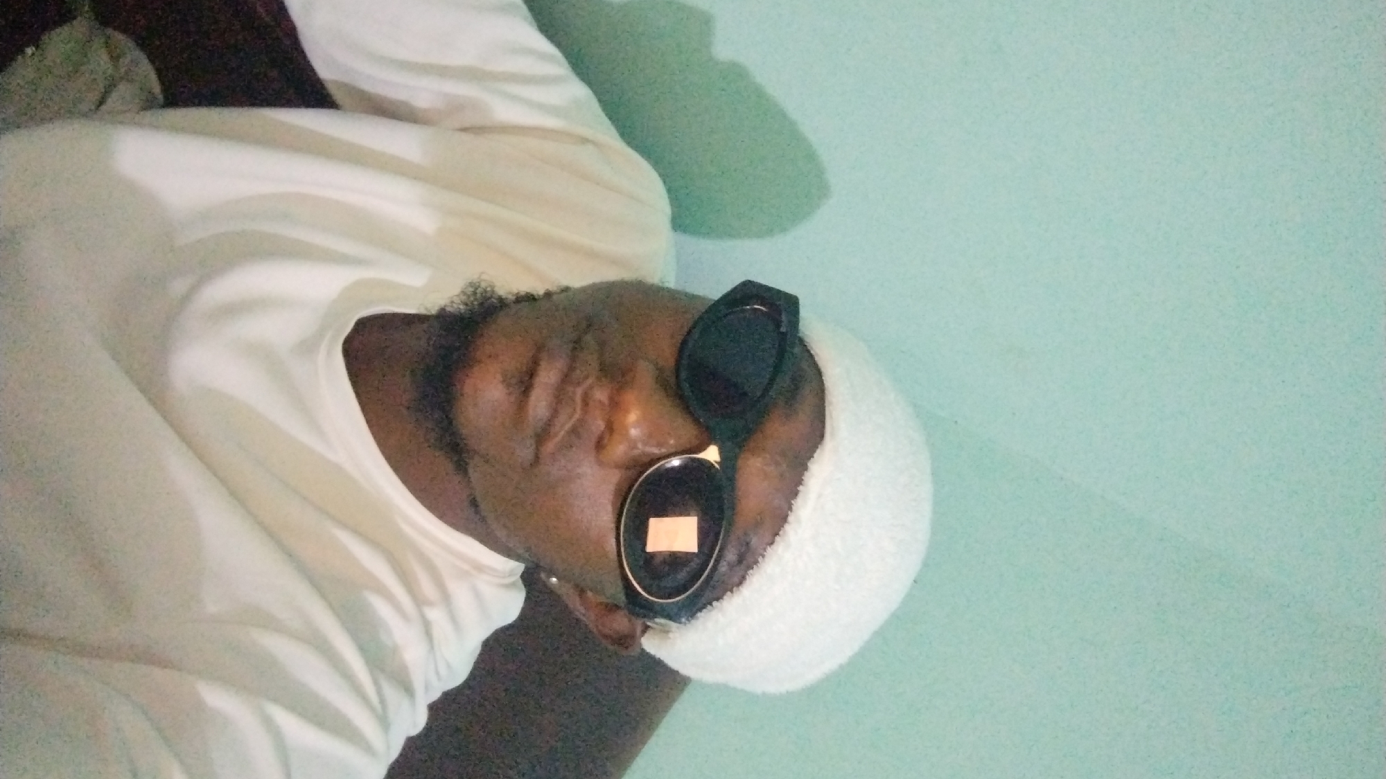 The image shows a close-up, selfie-style shot of a person wearing sunglasses and a white beanie. The person has a goatee and is wearing a cream-colored long-sleeved shirt. The background appears to be a wall painted a light teal color, with a shadow cast on it. There are no specific landmarks or indications in the image that would place the location in Jos, Nigeria, or any other city. The scene is indoors, and the lighting is dim, creating a shadow on the wall behind the subject.