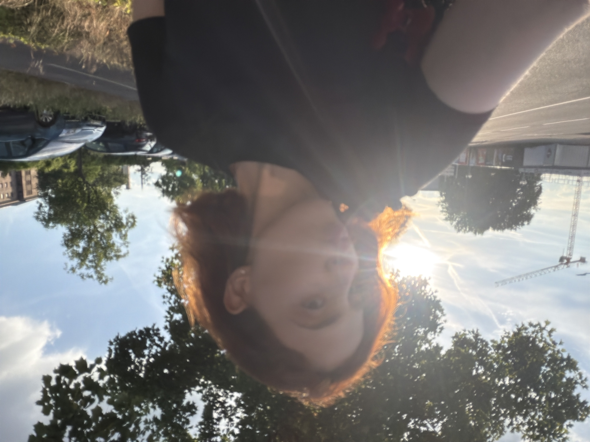 An outdoor, portrait-oriented photograph features a young person with reddish-brown hair, captured from the chest up, facing the camera. A strong lens flare from the sun, positioned behind and to the right of the subject, creates a radiant glow around their head and partially obscures their face. The person is wearing a dark-colored, likely black, short-sleeved top.
The background depicts an urban or suburban scene under a bright, sunny sky. Scattered white clouds and visible airplane contrails are present. To the left, several cars are parked alongside a road, with a multi-story building visible further in the distance. To the right, amidst a canopy of green trees, a large construction crane rises against the sky. The ground appears to be a paved surface or road. The bright sunlight suggests late afternoon or early evening. The setting is consistent with an urban environment like Strasbourg, France. No visible text is present.