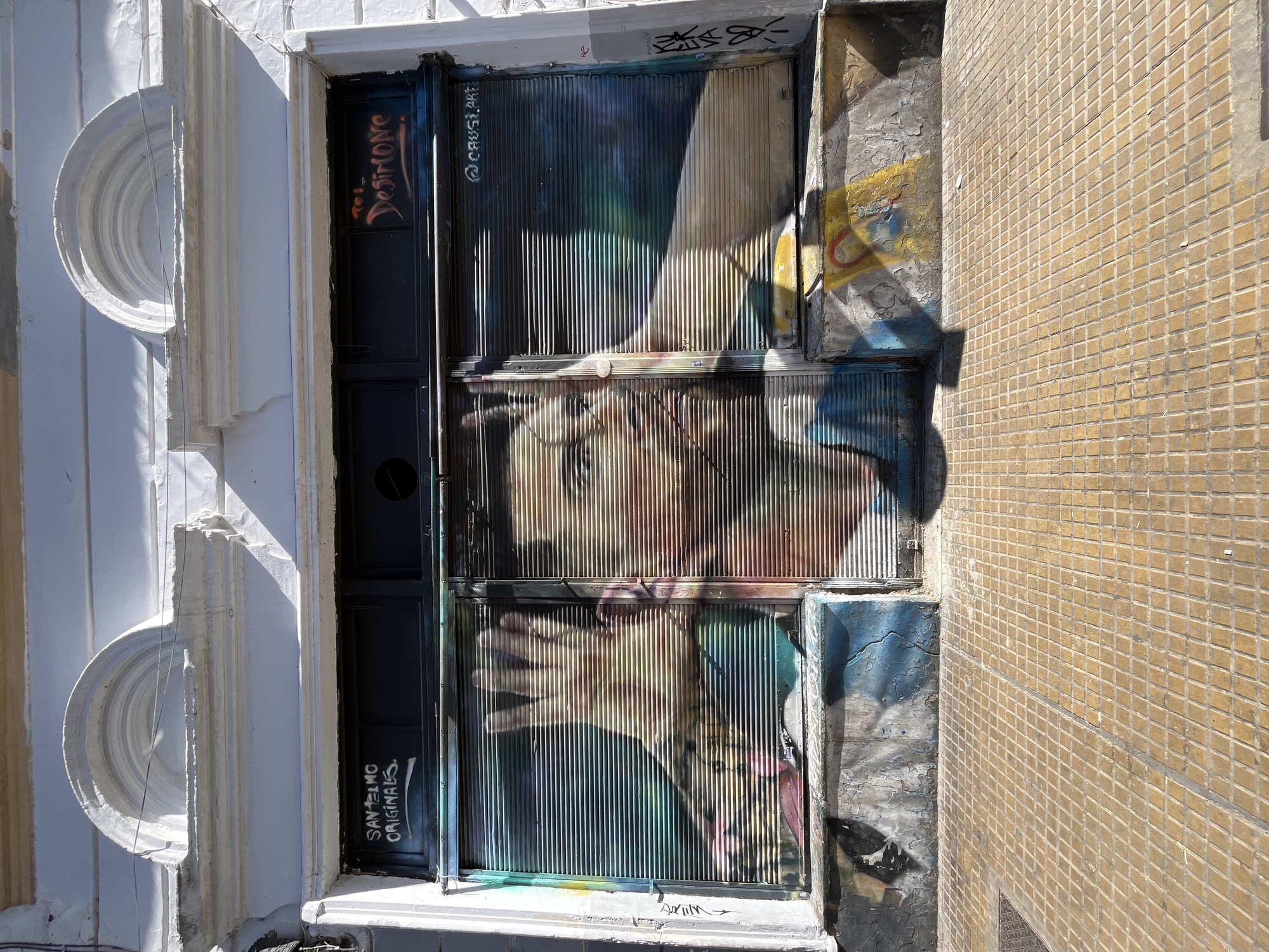 The image, oriented horizontally, captures an urban scene featuring a building facade, a shuttered entrance, and an adjacent tiled wall under bright sunlight, likely in Buenos Aires, Argentina.
On the left, a section of a white building facade is visible. It features two prominent circular, layered decorative elements, resembling recessed half-circles, stacked vertically. Below these, the facade has vertical fluting. Strong shadows are cast, highlighting the architectural details and suggesting a sunny day.
The central focus is a corrugated metal shutter or door, painted with a colorful mural. The mural depicts a stylized face, possibly a woman's, with prominent features and a discernible hand reaching across the lower part of the shutter. The corrugated texture of the surface distorts the artwork, creating a ribbed appearance. The color palette includes shades of blue, green, yellow, orange, and brown. Sunlight creates a diagonal shadow across the upper half of the mural.
Several pieces of graffiti are visible on and around the shutter. On a black panel to the left of the main mural, red-orange lettering reads "To: DESIRONE" at the top and "SAN TELMO ORIGINALS" in white at the bottom. On the upper left of the corrugated shutter itself, "@CAUSI.ART" is tagged in white. Other smaller, less distinct tags appear on the surrounding surfaces.
To the right of the shutter, a wall covered in small, light brown or tan square tiles extends vertically through the frame. The tiles are arranged in a regular grid pattern. The base of this tiled wall, near the shutter, shows signs of weathering and some faded graffiti, including yellow, blue, and red patches. Another small white tag, "WIIV" with an arrow, is visible at the very bottom center on a white ledge.
The overall impression is one of a vibrant street art installation set against older urban architecture, bathed in the strong light of a sunny day.
