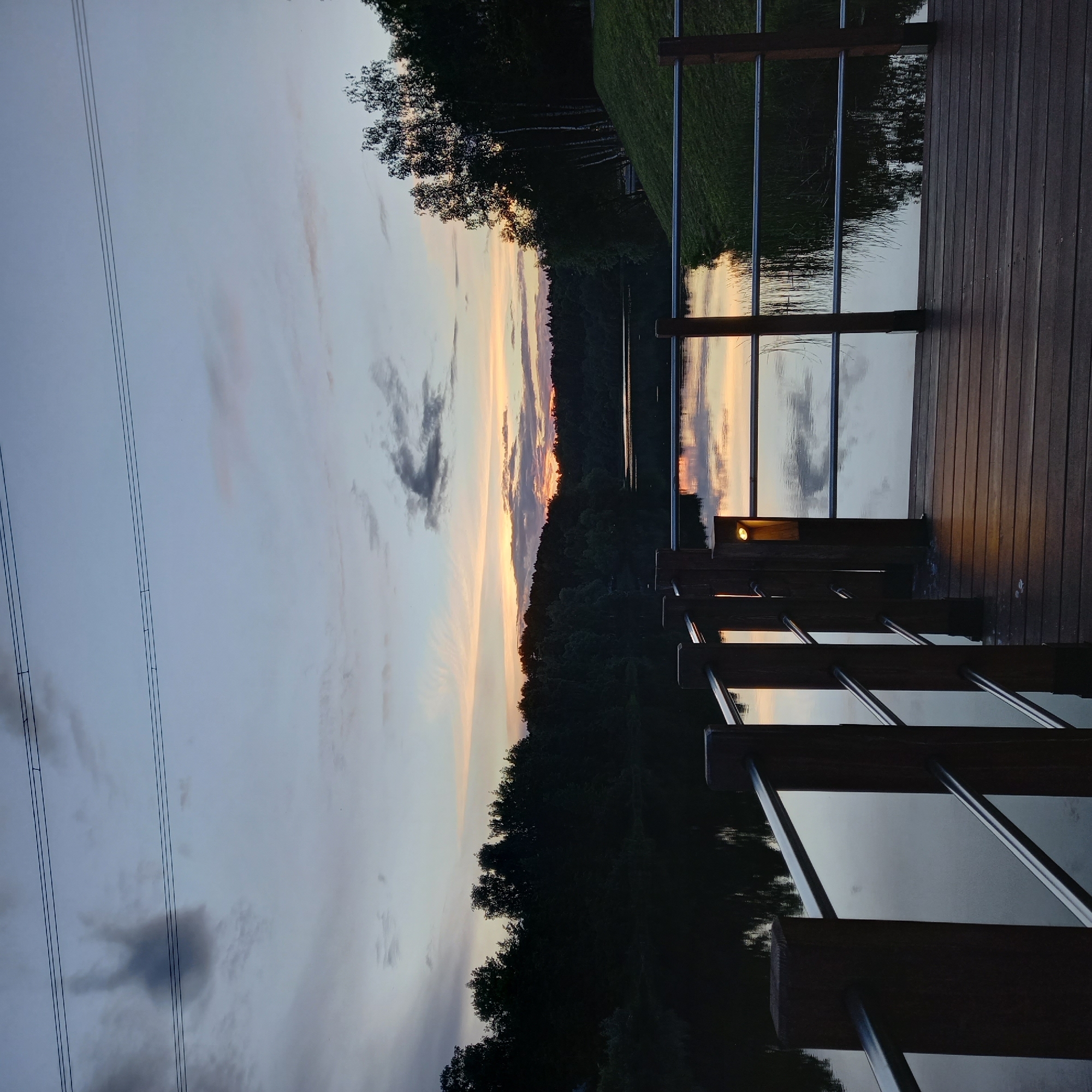 The image depicts a landscape at sunset, likely in a rural or suburban area. A body of water reflects the sky, which is filled with clouds tinged with orange and pink hues from the setting sun. The scene is framed by trees along the horizon, appearing as a dark silhouette against the bright sky.
On the right side of the image, there's a wooden deck or walkway with railings. A light fixture is visible on the deck, emitting a warm glow. High-voltage power lines are on the left side of the image.
Without additional information or visual cues pointing to Krasnogorsk, Russia, definitively identifying this location in Krasnogorsk based solely on this image isn't feasible. The generic features of water, trees, and sunset could apply to many locations.