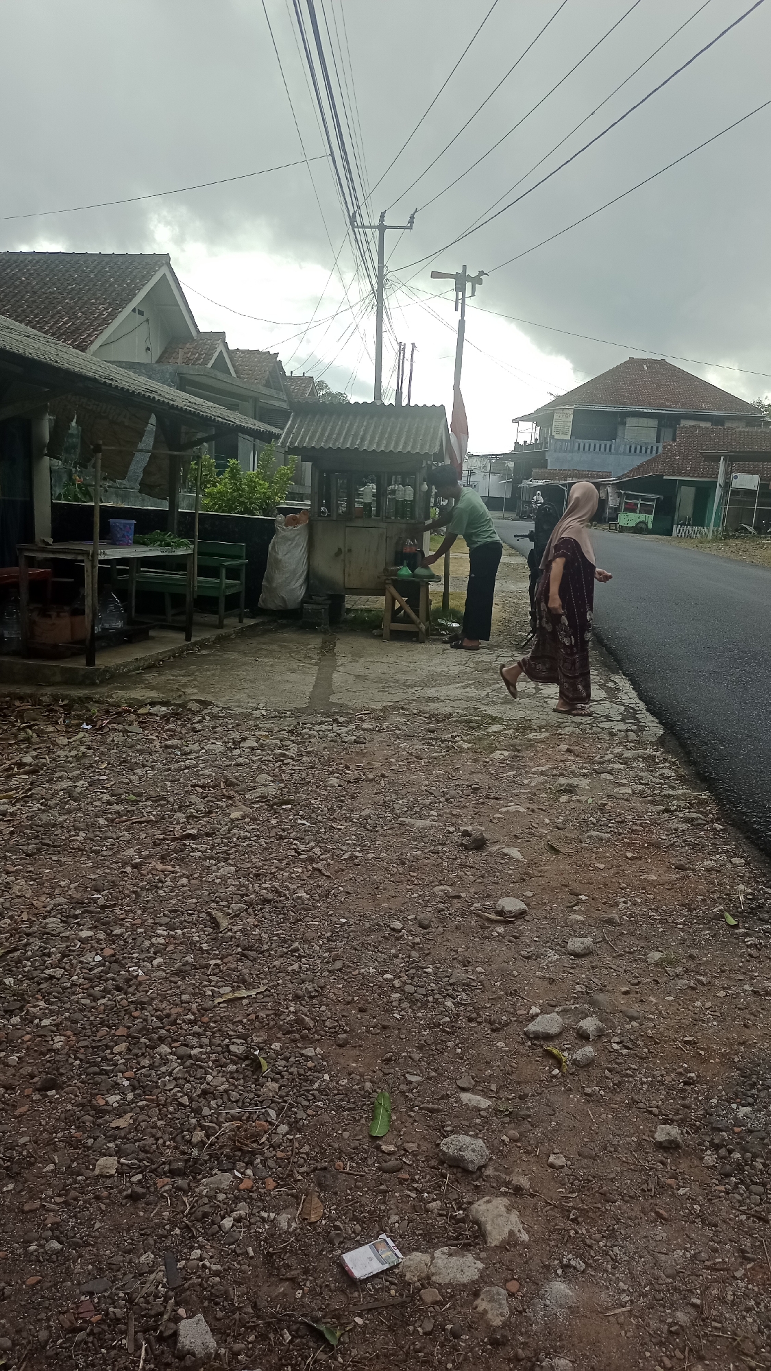 In the city of Karyamandala, Indonesia, a street scene unfolds under an overcast sky. The foreground is a gravel path leading to a paved road. On the left, a simple structure with a corrugated roof and open sides displays green benches and tables, suggesting a waiting or seating area. Next to it, a small, wheeled kiosk with a similar roof is attended by a person in a green shirt. They are leaning over the kiosk, seemingly interacting with its contents, with bottles visible. Further down the road, a person in a patterned dress and headscarf is walking away from the kiosk, toward the right side of the image. In the background, residential buildings with tiled roofs line the street, and a prominent utility pole with numerous wires crosses the sky. The overall atmosphere is that of a rural or semi-urban Indonesian street, with the kiosk appearing to be a small roadside vendor.