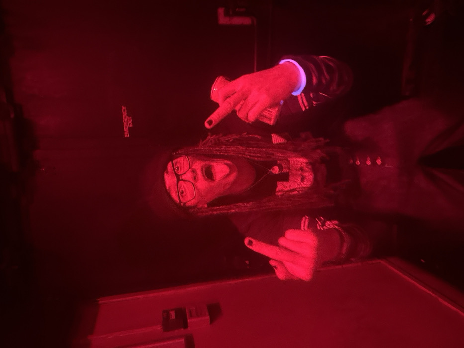 An individual with dreadlocks and glasses is captured in red lighting, sticking their tongue out and raising two middle fingers. One hand appears to hold a can or small object. The person wears a wristband that glows faintly. The background reveals a sign that says "Emergency Exit" and parts of what appears to be a door or wall.