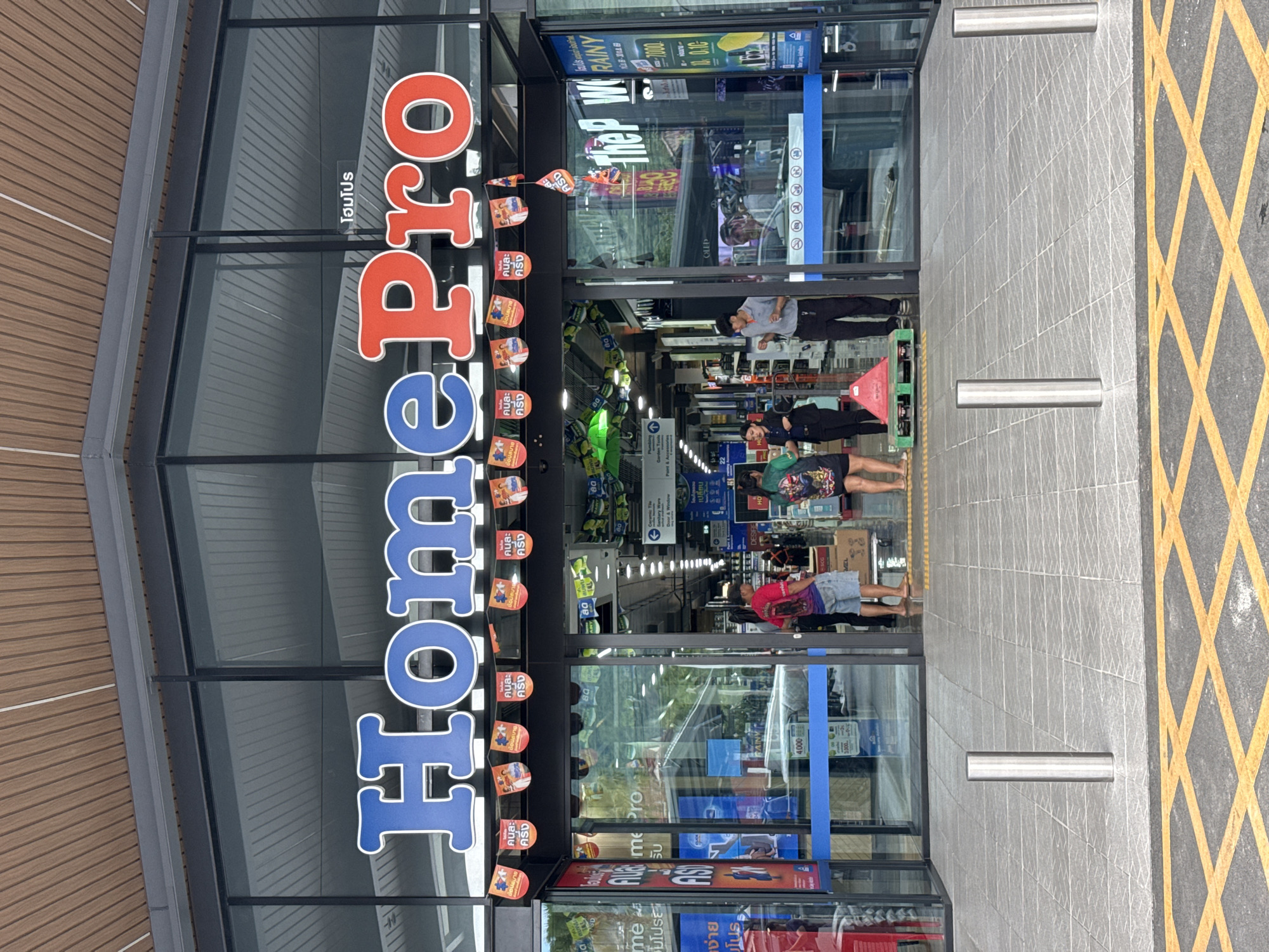 The image depicts the entrance of a HomePro store in Baan Chaweng Noi, Thailand. The prominent blue and red "HomePro" signage is visible above the glass doors. A small Thai sign, "โฮมโปร" (HomePro), is also present. Below the main sign, a string of small triangular banners with text and images hangs across the entrance.
Several people are visible at the entrance. On the left, a person in a red shirt and denim shorts stands facing away from the camera, with their hand reaching towards their pocket. To their right, a woman in a black top and skirt stands with her arms crossed. Further inside, a man in a grey shirt and black pants is seen speaking, holding what appears to be a small object in his hand. On the far right, two individuals are partially visible, one wearing a dark shirt and the other in a colorful patterned shirt and shorts.
The entrance area is paved with light-colored tiles. Metal bollards are positioned outside the entrance, and yellow cross-hatching is marked on the asphalt in front of the store. Inside the store, aisles of merchandise are visible, including shelving units and display items. The lighting inside appears to be a mix of overhead fluorescent lights and spotlights. A red and green pallet with a triangular red object on top is positioned near the entrance. Promotional signs and displays are scattered throughout the visible interior. The overall environment suggests a retail setting, likely a large home improvement store.