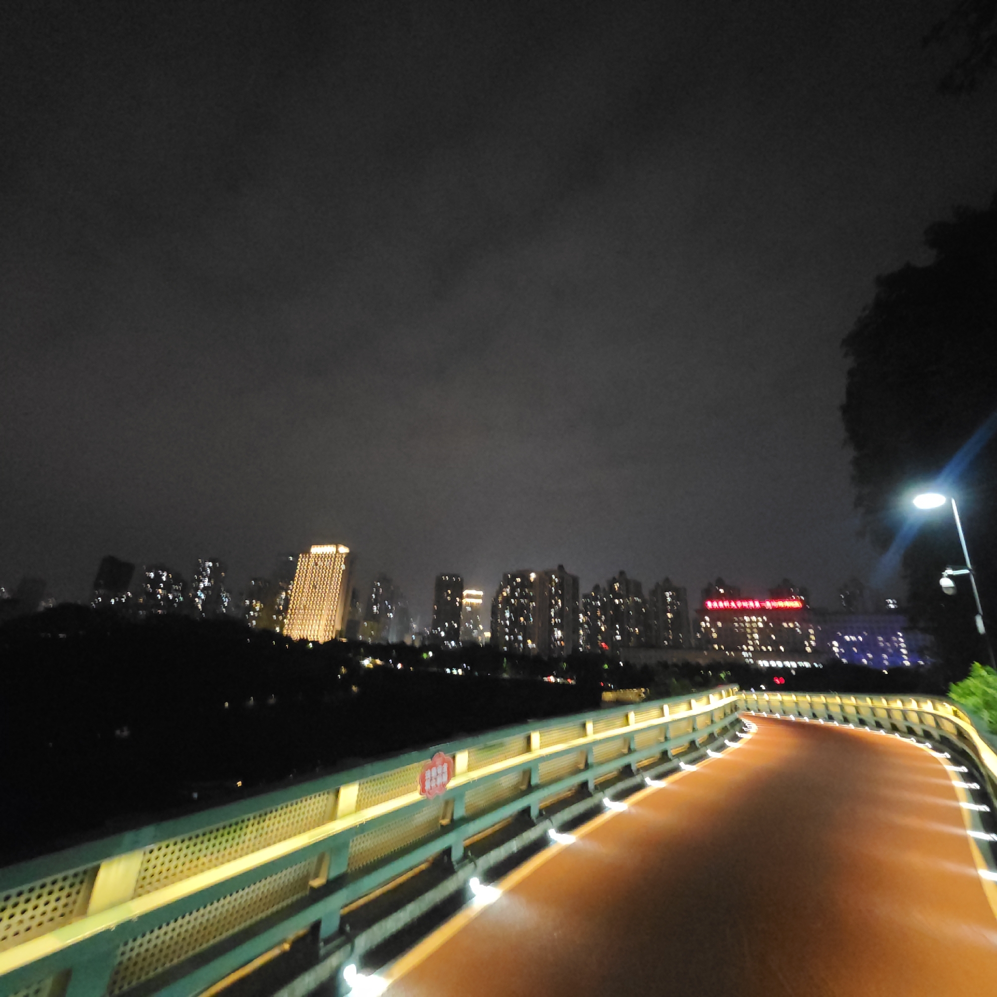 A nighttime shot depicts a curved, orange walking path flanked by green railings. Small white lights are embedded along the path's edges. Beyond the path, a row of dark trees separates it from a city skyline. Buildings are lit up with a variety of colors, primarily gold and white. A street lamp glows brightly to the right, partially obscured by a tree. The sky is overcast. The location is Yuzhong District, China.