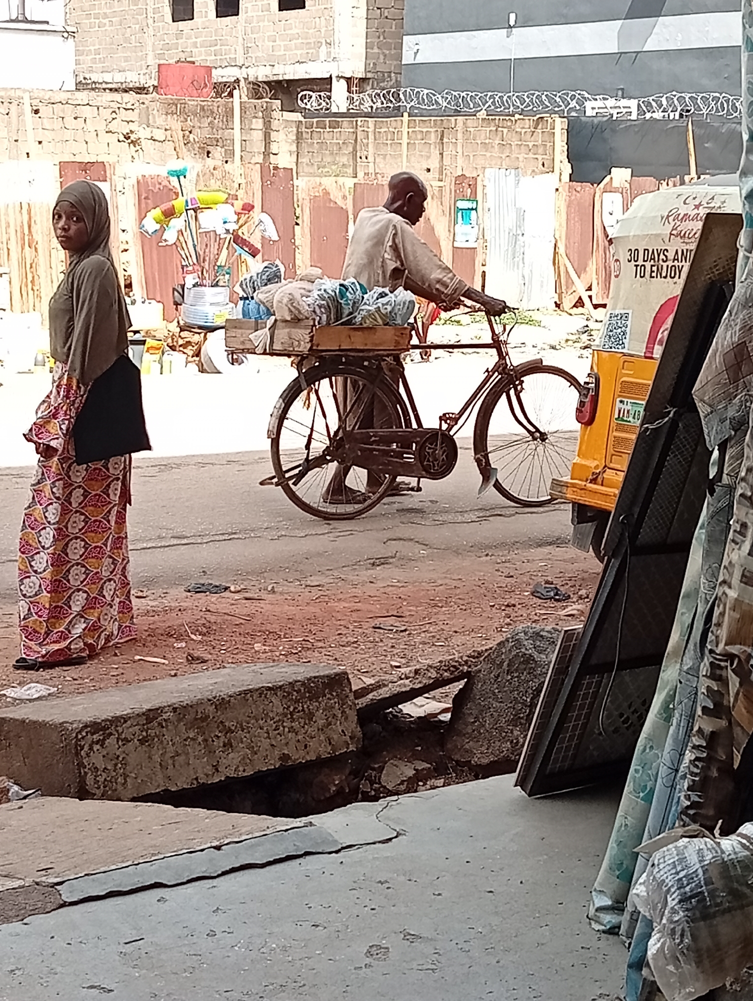 The image depicts a street scene in Maiburiji, Nigeria. A young woman wearing a hijab and a long, patterned skirt stands on the side of the road. A man is riding a bicycle with a wooden crate mounted on the back, filled with what appears to be bundled textiles or fabrics. A yellow auto-rickshaw is partially visible behind the bicycle. The background includes buildings with corrugated metal fencing topped with barbed wire. Various items, possibly merchandise, are displayed near the building in the background. The foreground shows a broken curb and scattered debris, suggesting an environment in need of repair.