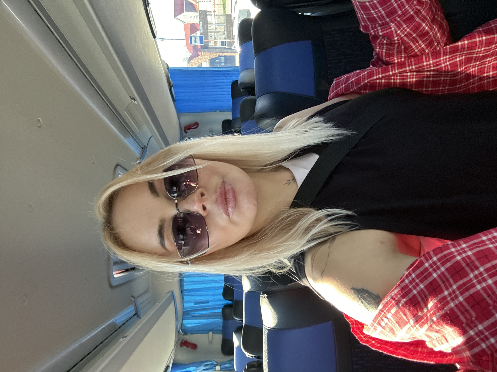 A woman with long blonde hair and sunglasses is seated on a bus with blue seats. She wears a black shirt, a red and white plaid shirt, and a seatbelt. Outside the window is a building with a sign in Russian, indicating the location is Megion, Russia.