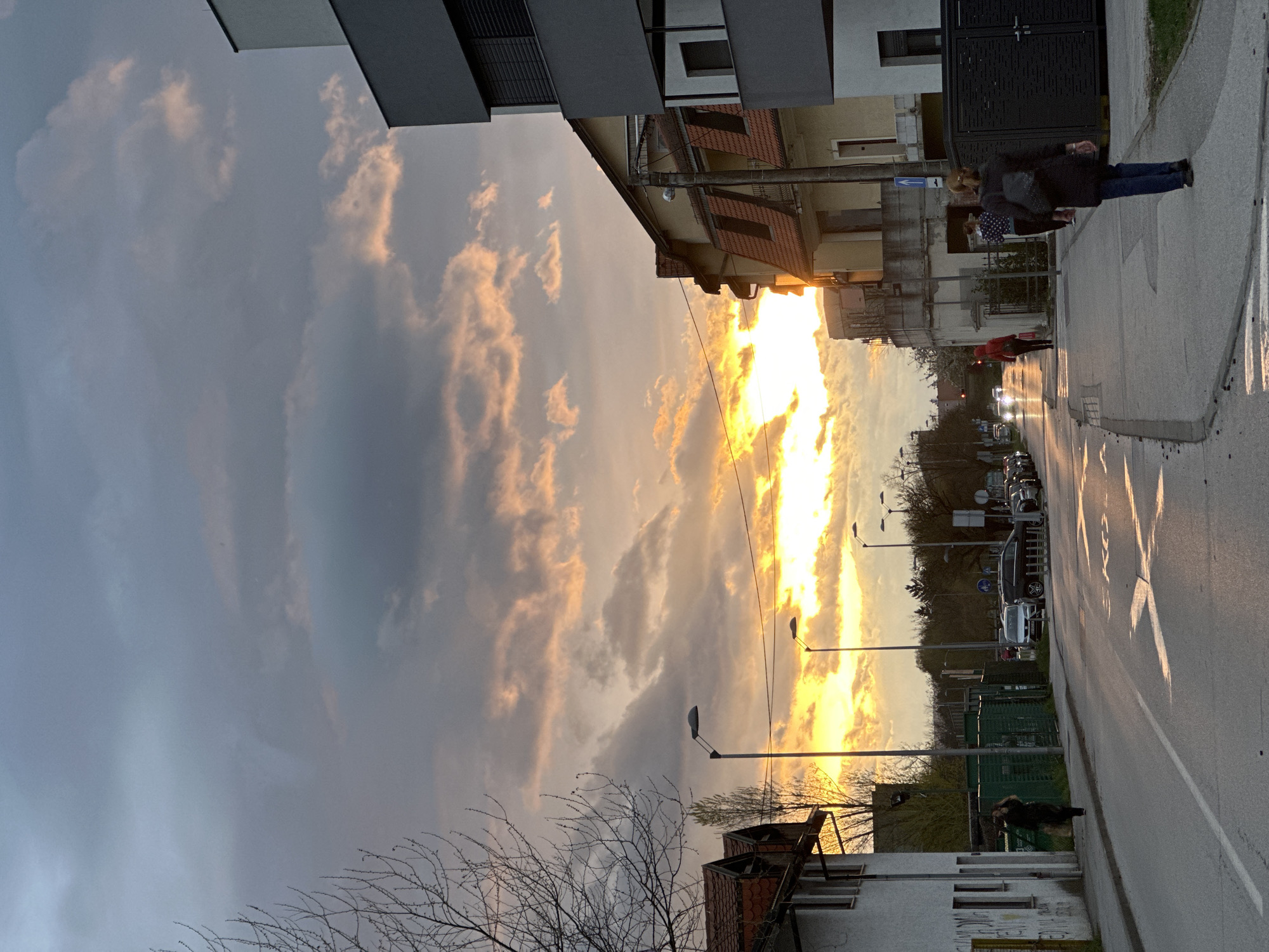 The image depicts a street scene in Zagreb, Croatia at sunset. The sky is filled with clouds illuminated by the setting sun. Buildings line both sides of the street. People are walking on the sidewalk. Cars are parked on the side of the road. A lamppost is visible. Trees are in the background.