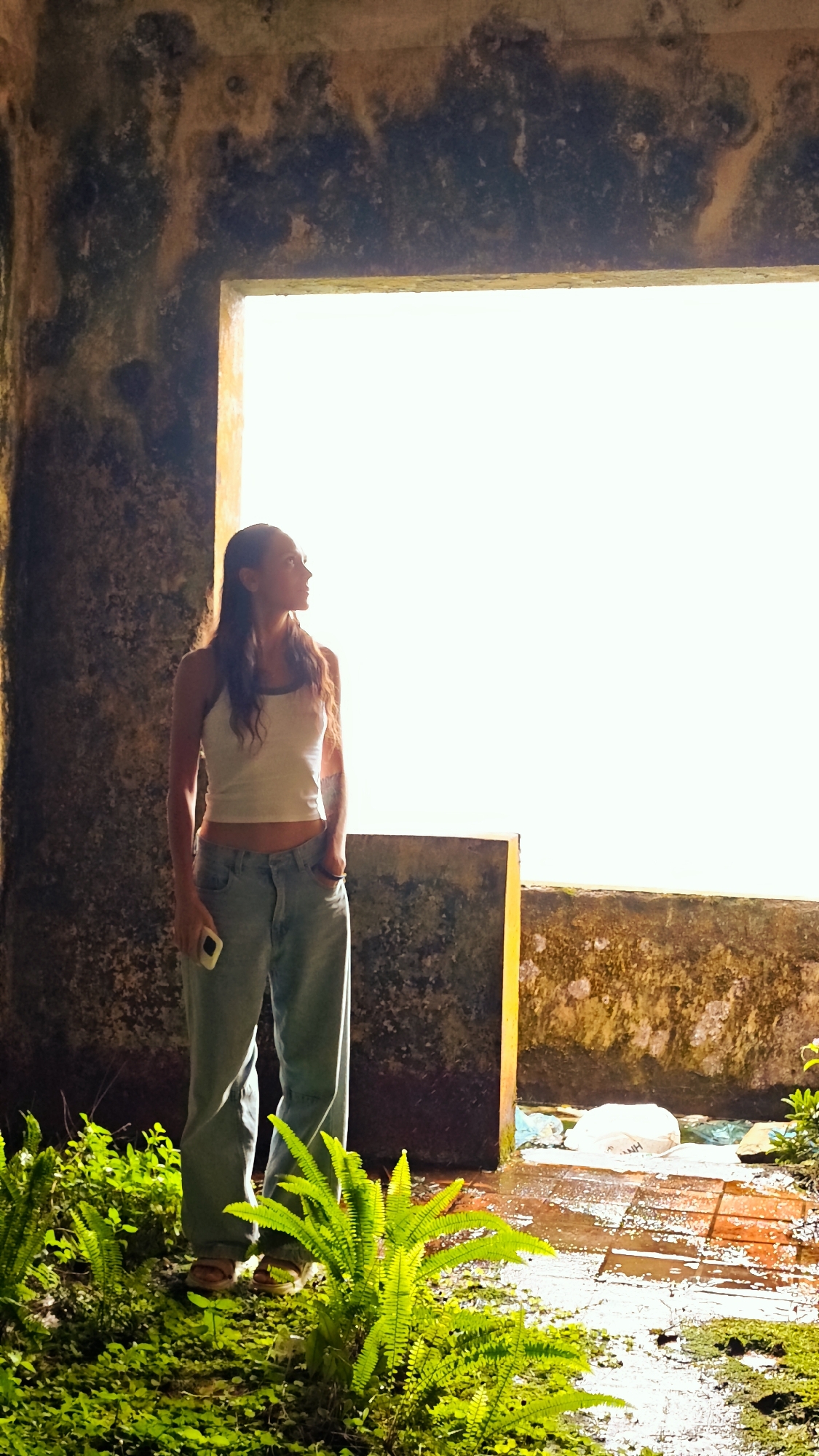A woman stands in a dimly lit, stone structure with a large, bright opening to her right, looking out. She is wearing a white tank top and blue jeans, with her right hand in her pocket and her left hand holding a light-colored smartphone. Lush green ferns and foliage grow in the foreground, with wet, reddish-brown brick flooring visible. The walls of the structure are rough and weathered, with patches of dark and light discoloration. The bright opening suggests a large window or doorway leading to an outdoor area. The overall impression is one of contemplation and a stark contrast between the enclosed, aged interior and the bright, possibly natural, exterior. The location is specified as Mẫu Sơn, Vietnam.