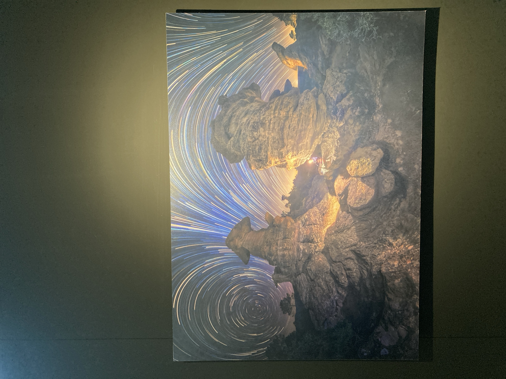 The image shows a vertical rectangular print mounted on a dark, matte wall. The print features a night-time landscape with star trails circling a rocky terrain. The rocks are illuminated with a warm light, suggesting a light source within the cave. The star trails are concentric circles of light, indicating a long exposure photograph with the camera fixed on a point in the sky. The location is unknown.