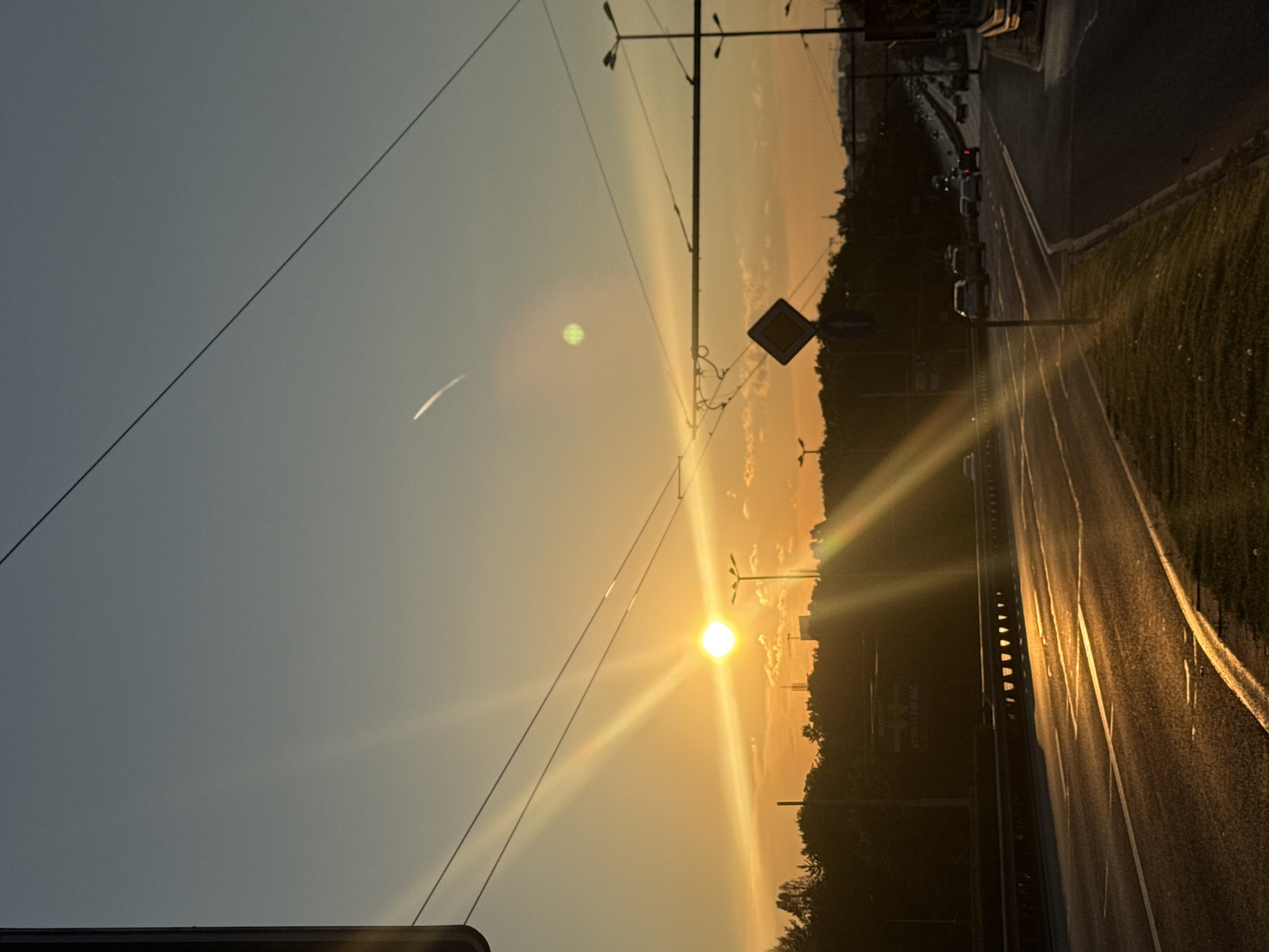 This image depicts a sunset on a highway in Sofia, Bulgaria. The sun is low on the horizon, casting a warm, golden light across the scene. The sky is a gradient of orange and blue, with a few wispy clouds. There are power lines crisscrossing the sky, and a traffic sign is visible on the right side of the road. Cars are driving on the highway, and trees line the sides of the road. The overall impression is one of peaceful twilight in an urban setting.
