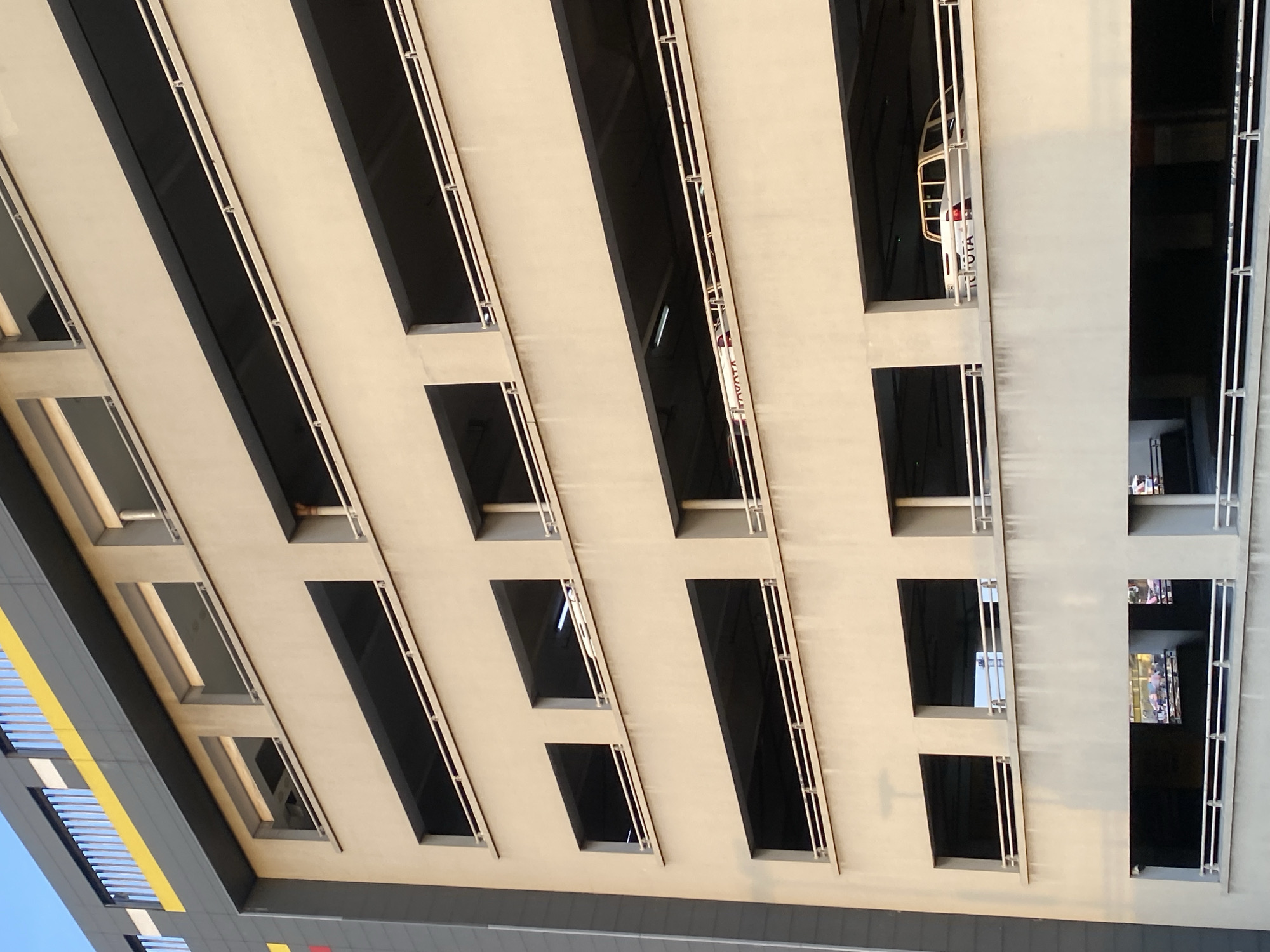 The image shows the exterior of a multi-level parking garage, presumably in Falomo, Nigeria. The structure is light-colored, with repeating rectangular openings that are likely parking spaces. Dark recesses indicate the interior of the parking levels. Metal railings are visible along the outer edges of the openings. On one level, a white Toyota car is visible inside one of the parking spaces. The building’s design includes a section with yellow and gray horizontal accents. The photograph is taken from a low angle, looking upwards. There are at least 2 humans visible.