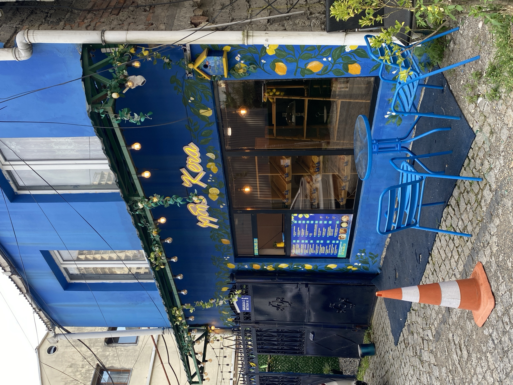 The image depicts a blue-colored building with a street-facing storefront decorated with lemon-themed murals. The storefront features a large window displaying baked goods and a menu board. In front of the store, there is a small table with two blue chairs, a traffic cone, and a black metal gate. The street is paved with cobblestones, and the overall setting suggests an urban environment, likely a commercial establishment. A sign on the gate indicates "METAXA st. 6". The style of the building and the surrounding architecture are consistent with the city of Batumi, Georgia.