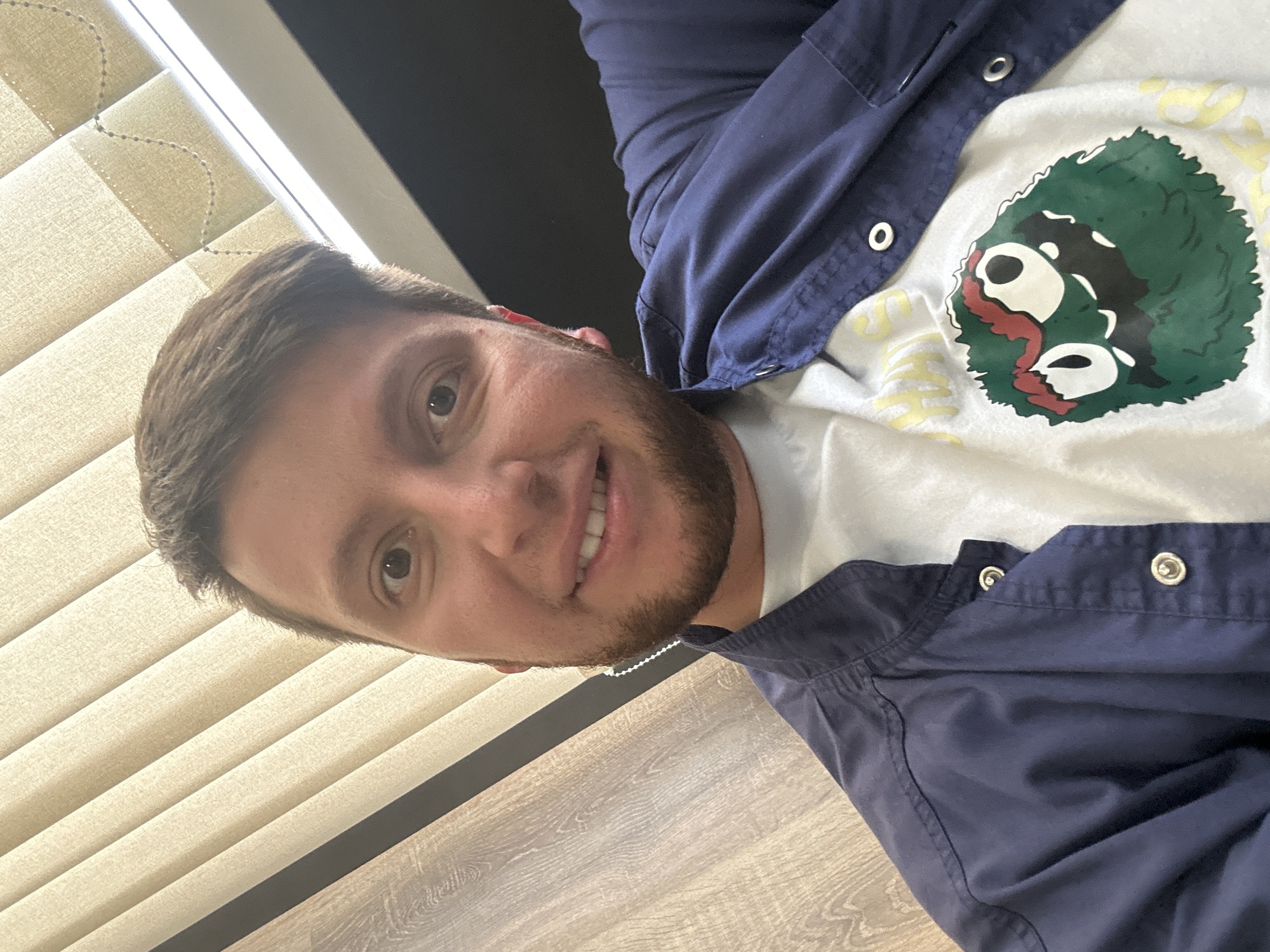 The image is a close-up, high-angle selfie of a young man with short brown hair and a beard, smiling. He is wearing a dark blue collared shirt unbuttoned over a white t-shirt with a cartoonish green monster graphic. The background appears to be an office environment with vertical blinds and a wooden-textured surface. There is no indication of the location in Moscow, Russia.