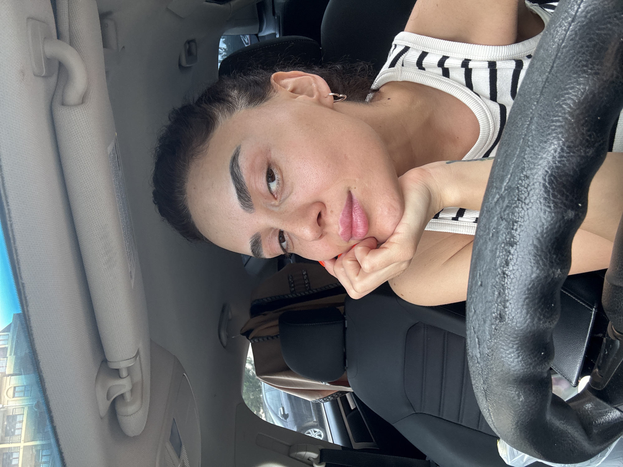 A young woman with dark hair pulled back is seated in the driver's seat of a car, her right hand propping up her chin. She is looking directly at the camera, her lips slightly pursed. She is wearing a black and white striped tank top. The car's interior is visible, including the steering wheel, dashboard, and the interior of the roof with a sun visor. A light brown jacket with patterned trim is hanging from a hook on the back of the passenger seat. Outside the car window, another dark-colored car is parked. The image is taken during the daytime, with natural light illuminating the scene. No specific landmarks in Tbilisi, Georgia are identifiable in this image.