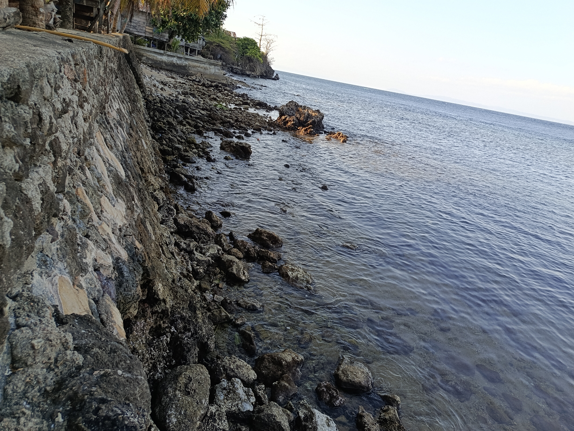 The image depicts a coastal scene at the edge of Mansalean, Indonesia. A rough, grey stone wall forms the foreground, leading to a rocky shoreline scattered with various-sized stones and boulders. The clear, blue water of the sea gently laps against the shore, with some submerged rocks visible beneath the surface. In the background, a vegetated slope rises, with a few structures and palm trees visible, suggesting a rural or semi-developed area. The horizon is marked by a faint line of distant land against a clear, pale blue sky.