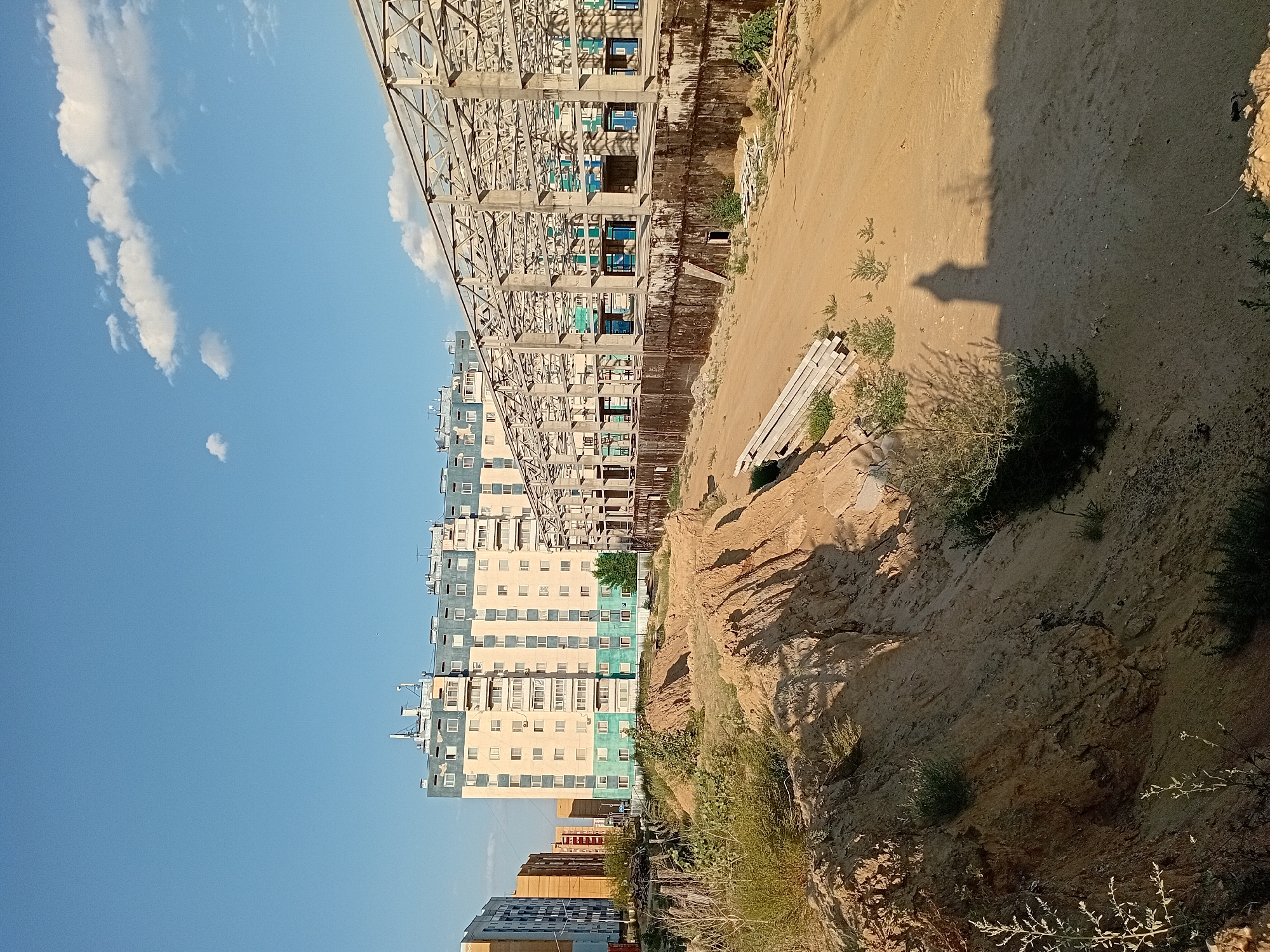 The image captures a construction site in Semey, Kazakhstan, characterized by a clear blue sky with scattered white clouds. In the background, several multi-story apartment buildings stand, one particularly prominent with light yellow and teal facade and multiple balconies. Adjacent to these residential buildings, a large, unfinished structure with a metal frame and concrete supports is visible, suggesting ongoing construction. In the foreground, a sandy terrain with uneven, excavated earth mounds and sparse vegetation dominates the scene. A shadow of a building, possibly the unfinished structure, is cast across the sandy ground. A pile of concrete slabs lies on the left side of the frame, and other construction debris is scattered throughout the area. The overall impression is that of a developing urban landscape with active construction amidst existing residential areas.
