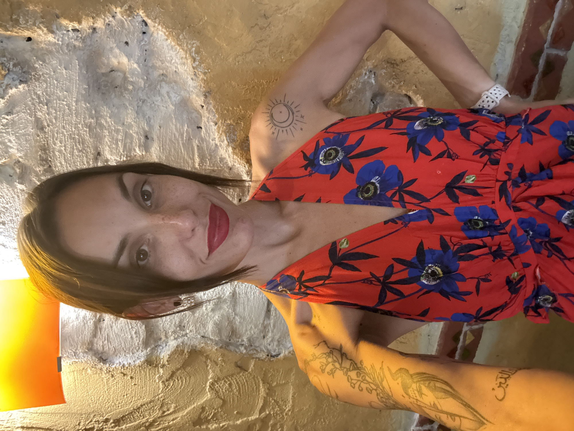 A woman with dark brown hair and fair skin with freckles is pictured. She is wearing a red dress with a blue floral pattern. She has a tattoo of a bird on her left arm and a sun and moon tattoo on her right shoulder. Her eyes are brown, and she is wearing red lipstick. She is smiling and has her left hand on her hip. A light fixture with an orange glow is visible in the upper left corner of the image. The background appears to be a textured, light-colored wall. The woman is standing indoors, possibly in a restaurant or bar, in Pleven, Bulgaria.