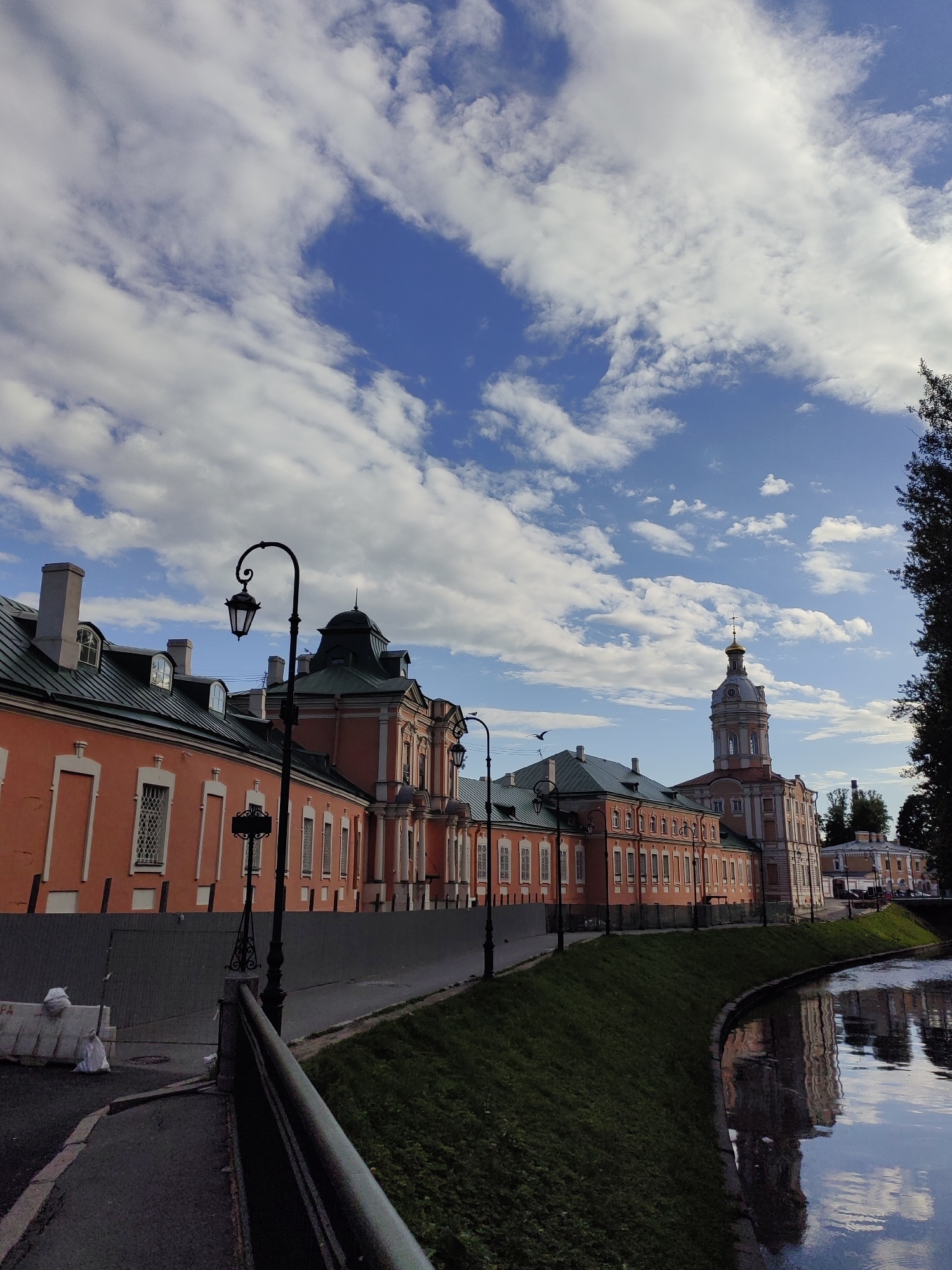 The image depicts a picturesque canal-side view of historical buildings in Saint Petersburg, Russia. The architecture is characterized by ornate facades, predominantly in a warm, salmon-pink hue, with contrasting green roofs. Several windows with intricate white detailing are visible along the buildings. A tall, slender bell tower with a golden dome and cross stands prominently, adding to the grandeur of the scene. The canal, reflecting the sky and buildings, flows gently alongside a grassy embankment.
Several decorative lampposts line the walkway that runs along the canal's edge. The sky is a vibrant blue, dotted with fluffy white clouds, suggesting fair weather, possibly late afternoon or early evening given the soft light and the reflections on the water. The scene evokes a sense of tranquility and historical charm. There is no visible activity of people, and the focus is on the architecture and the natural elements of the sky and water. The presence of a construction barrier and some plastic bags on the left side of the image suggests ongoing urban work.