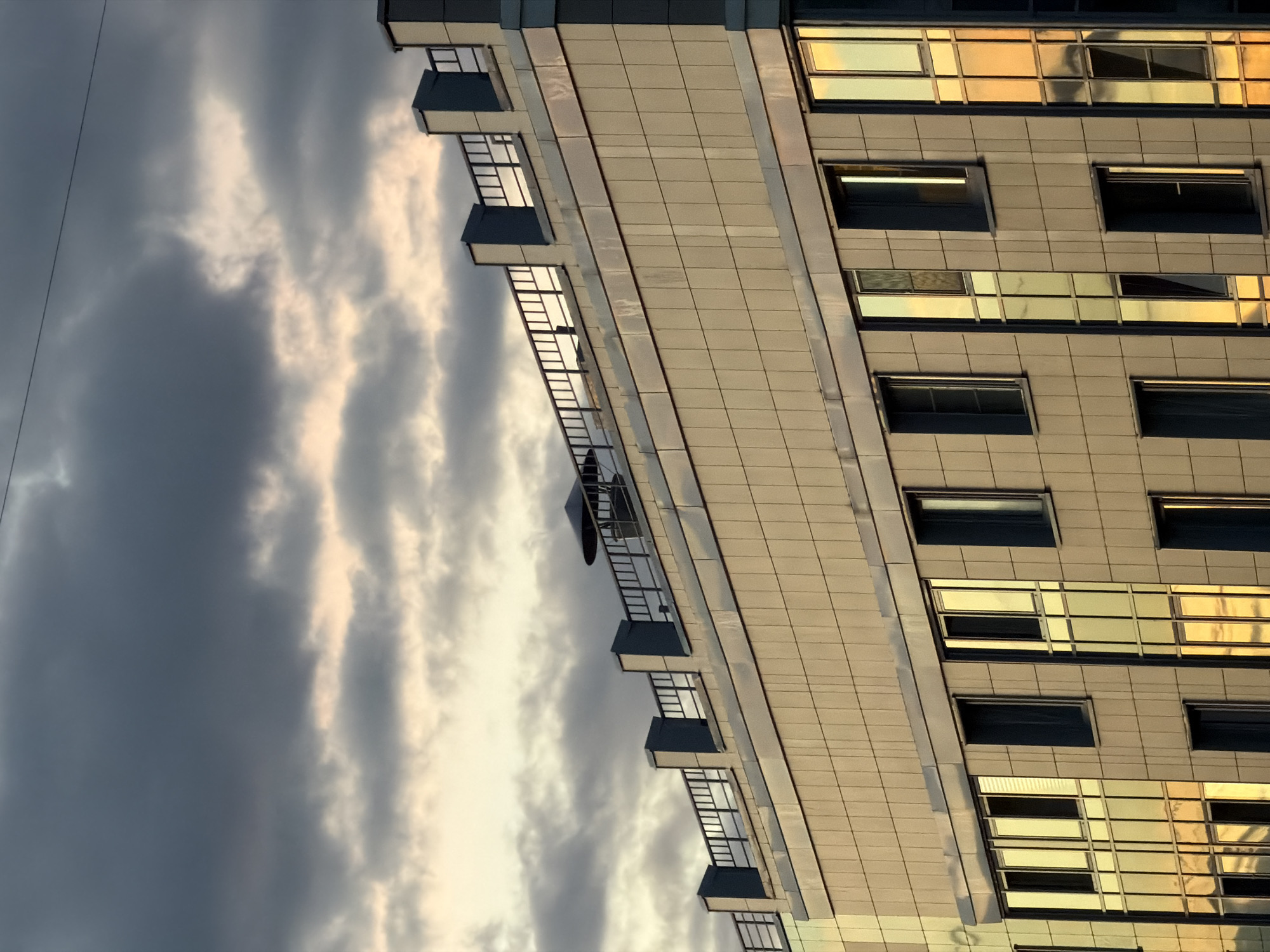 The image shows a close-up, low-angle view of the upper floors and rooftop of a modern building. The building's facade is clad in light-colored, square tiles, with horizontal bands of a darker material at intervals. Numerous windows are visible, many of which are reflective and capture the golden hues of the sunset and the grey, cloudy sky above. A dark railing runs along the top of the building, supported by square pillars. A ventilation unit is also present on the roof. The sky is filled with dramatic, dark grey clouds, with patches of lighter, illuminated cloud near the horizon. The overall scene suggests an urban environment during the late afternoon or early evening.