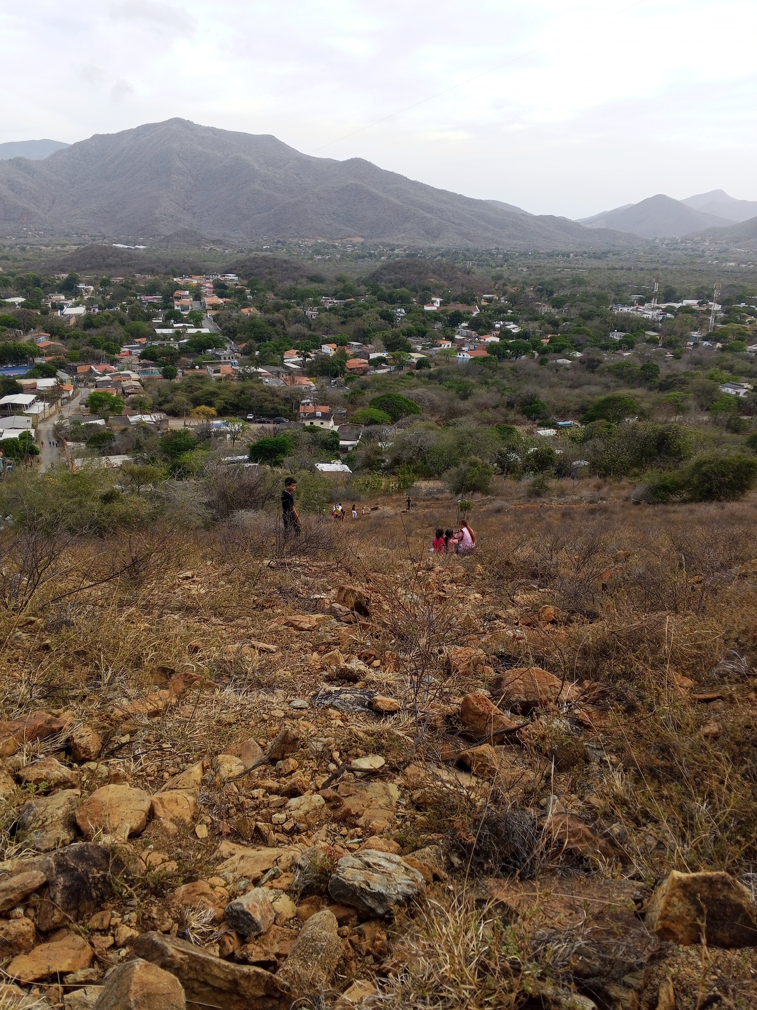 The image depicts a rocky, dry hillside overlooking a town situated in a valley surrounded by mountains. The foreground is dominated by a rough terrain of reddish-brown rocks and dry vegetation. Further up the hill, there's a group of people; one appears to be a taller individual walking down, while another, smaller group seems to be sitting or standing. In the middle ground, there's a sprawling town with houses and buildings interspersed with green trees. The town is nestled in a valley that stretches towards the base of the mountains in the background. The mountains are gray and appear dry, with a hazy sky above them.