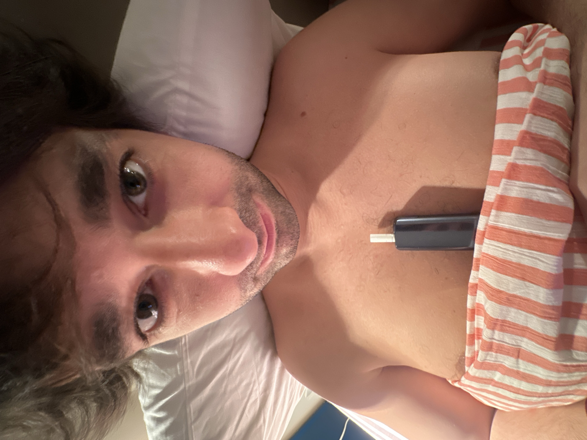 The image is a close-up, eye-level shot of a person lying in bed. The person is facing the camera, and their head is propped up on a white pillow. Their dark, curly hair is visible around their head. The person has brown eyes and a light beard. They are shirtless, and a striped blanket is covering their lower chest and abdomen. A small, dark electronic device with a white tip is placed vertically on their chest, above the blanket. The lighting is warm and appears to be artificial. There is no visible indication of the location being Moscow, Russia.