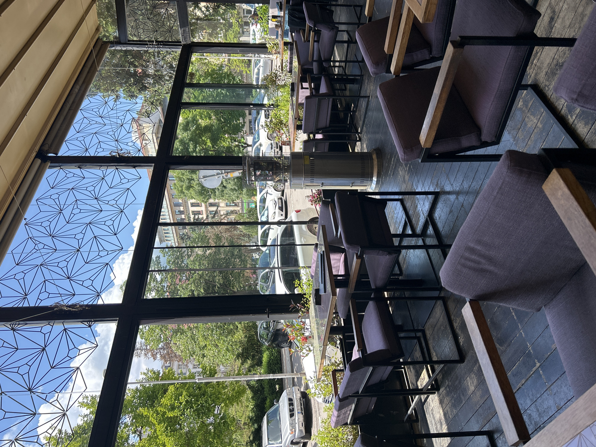 The image shows an indoor cafe space, likely during the day. The furniture consists of dark wood tables and purple upholstered chairs. Large windows offer an exterior view of a street lined with trees, buildings, and parked cars. A patio heater is visible near the windows. The ceiling is covered with triangular geometric patterns. The location appears to be Unknown, Unknown.