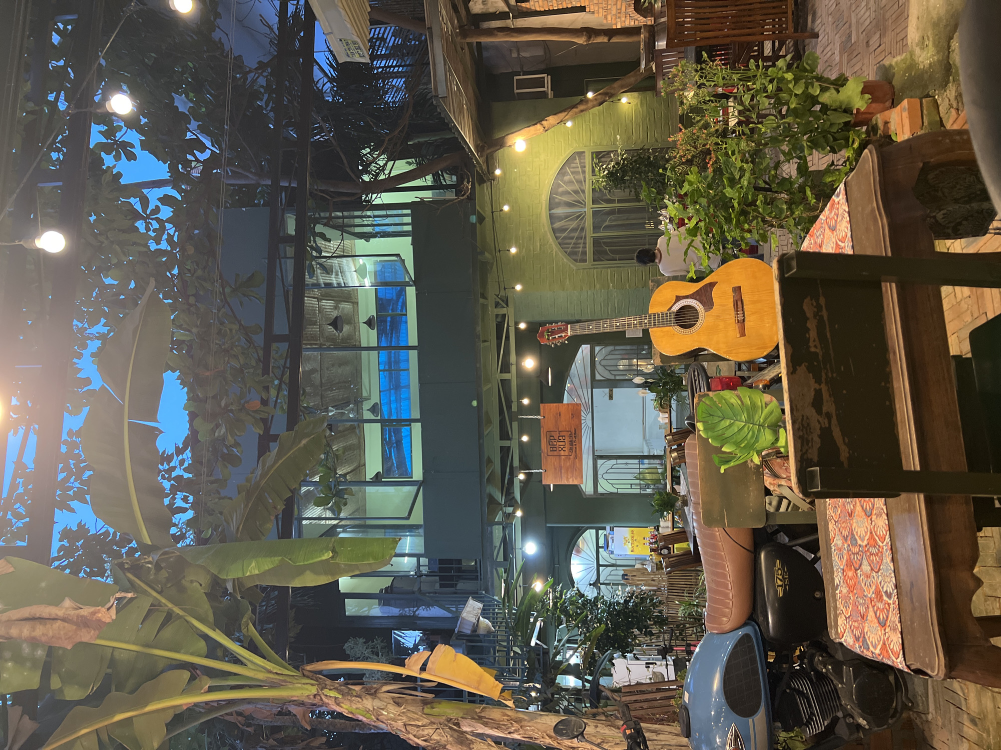The image captures the outdoor seating area of a cafe in Nha Trang, Vietnam. The seating area is surrounded by lush greenery, including banana trees. Overhead, there are string lights adding to the ambiance. A building with large glass windows is visible in the background. A guitar rests on a table next to a vintage motorcycle. A man in a white shirt is sitting near the guitar and motorcycle.
