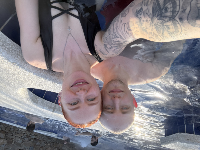 The image captures a selfie of a couple in a hot tub or heated swimming pool, situated in Rostov-on-Don, Russia. The male subject on the left, with very short hair, fair skin, and blue eyes, is partially submerged in the dark blue water, looking towards the camera with a slight smile. His left arm, visible in the foreground, is adorned with extensive black-and-white tattoos, including a large butterfly motif. The female subject on the right, with red hair, fair skin, and blue-green eyes, smiles broadly at the camera. She presents multiple facial piercings (nose ring, septum ring, lip ring, and ear piercings) and wears a black one-piece swimsuit featuring a criss-cross detail at the cleavage, complemented by a silver chain with a crucifix pendant. Her right arm extends forward, presumably holding the camera. In the background, three to four additional individuals, predominantly males, are partially visible in the steaming water. The pool features a light-colored, textured edge and dark blue tiling. A natural stone wall is visible beyond the pool structure. The scene is illuminated by bright sunlight, reflecting prominently off the water's surface.