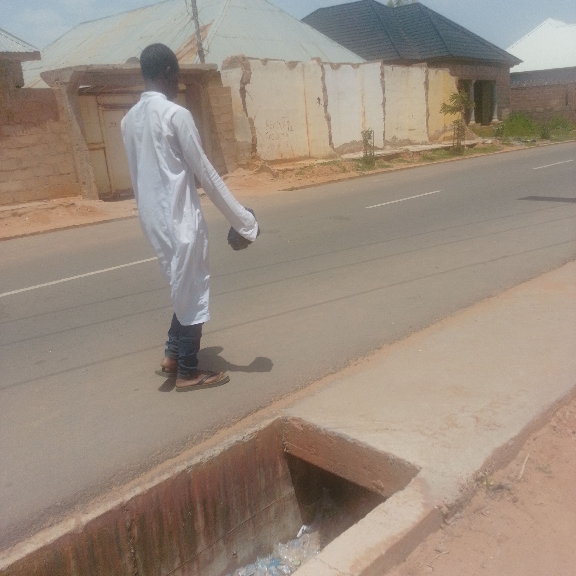 A young man in a white kaftan and dark trousers stands on the side of a paved road, holding a dark object in his right hand. He is wearing flip-flops. In the foreground, there is a drainage ditch filled with plastic bottles. Behind him, there are buildings with corrugated metal roofs and some with crumbling walls. The scene appears to be in a semi-urban area. The sky is bright, suggesting it is daytime, and the lighting indicates fair weather. There is some graffiti on one of the walls, but it is not clearly legible. The man's posture suggests he might be walking or paused on his journey.