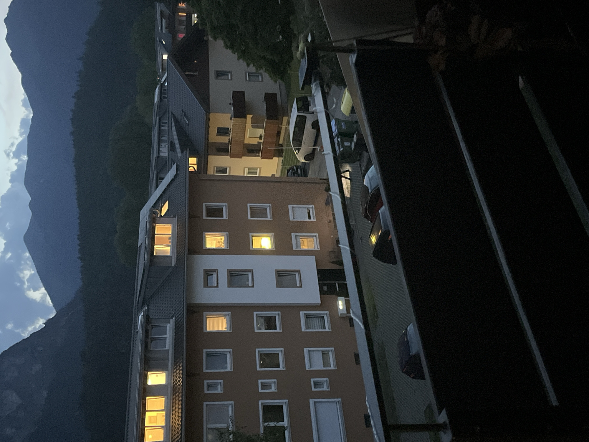The image depicts a residential area at dusk in Garmisch-Partenkirchen, Germany. Several apartment buildings with lit windows are visible, suggesting indoor occupancy. In the foreground, a dark railing or balcony separates the viewer from the scene. Cars are parked in a lot, including a white van and other dark-colored vehicles. Further in the background, dense evergreen trees line the base of dark, imposing mountains that dominate the skyline under a cloudy sky. The overall atmosphere is one of quiet evening in a mountainous town.