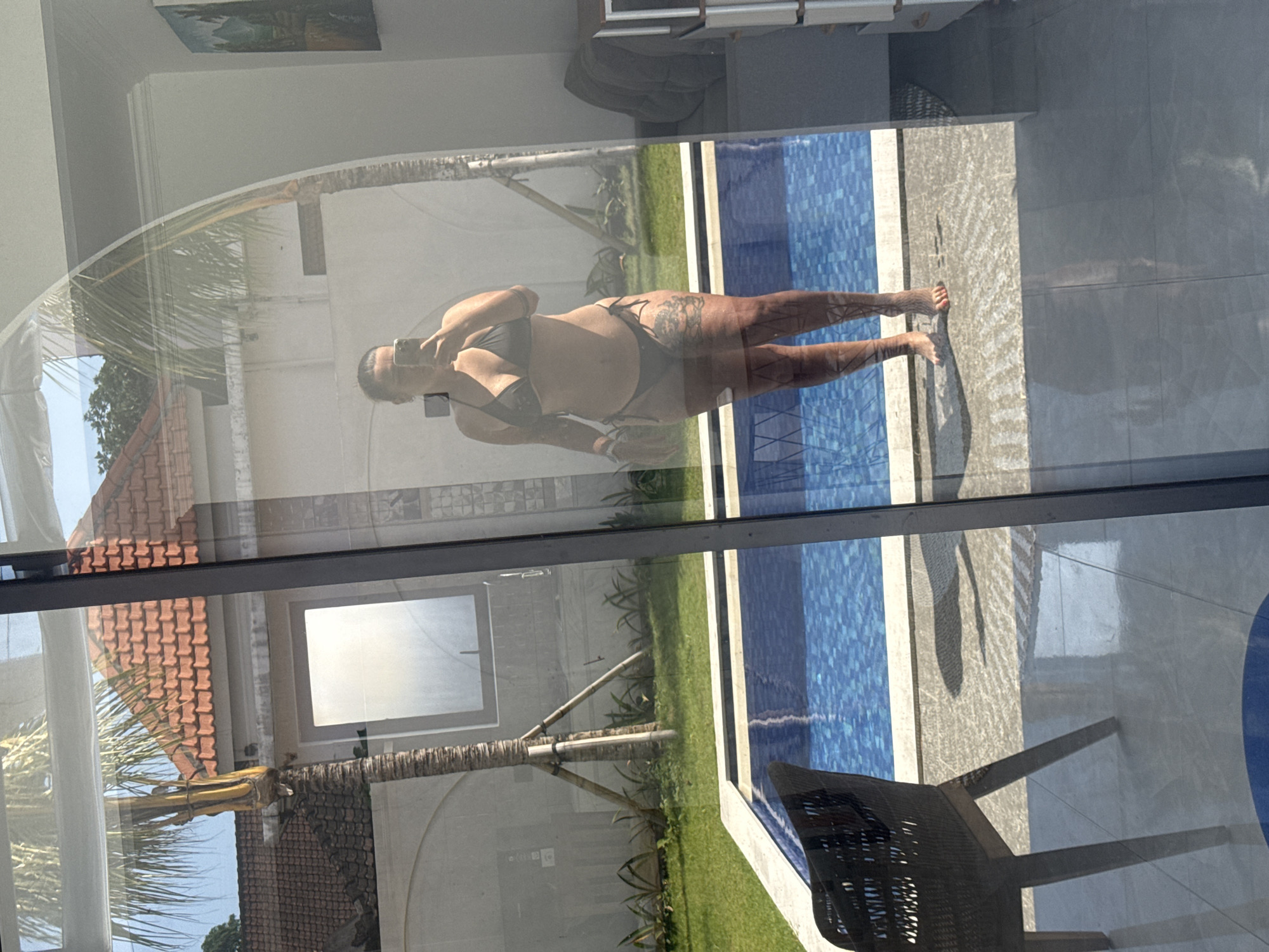 The image displays a woman's reflection in a large glass door, revealing an outdoor pool area and traditional buildings, set in North Kuta, Indonesia. The woman, standing barefoot on a light-colored patio next to a swimming pool, faces the glass and holds a dark-colored smartphone to take a selfie. She wears a black two-piece bikini, featuring a triangle top and tie-side bottoms, and has visible tattoos on her legs and thighs.
Outside the glass, beyond the pool, is a meticulously maintained garden with green grass and several tropical plants. A large palm tree with multiple support struts is centrally located in the garden. In the background, several buildings with distinctive red-tiled roofs and light-colored walls are visible under a clear, bright sky. One building features a prominent frosted rectangular window. The rectangular swimming pool has blue mosaic tiles and a white coping.
Reflected on the glass from the interior, parts of a dark brown woven chair with wooden legs are visible on the left, positioned on the patio outside. On the right, the reflection shows a dark gray cushioned furniture piece, possibly a chair or sofa back, and a light-colored cabinet or chest of drawers. A framed piece of art or a mirror is also faintly reflected on the upper right wall. The interior floor visible through the bottom of the glass appears to be dark gray tiled. Bright sunlight casts shadows on the patio, particularly behind the woman.