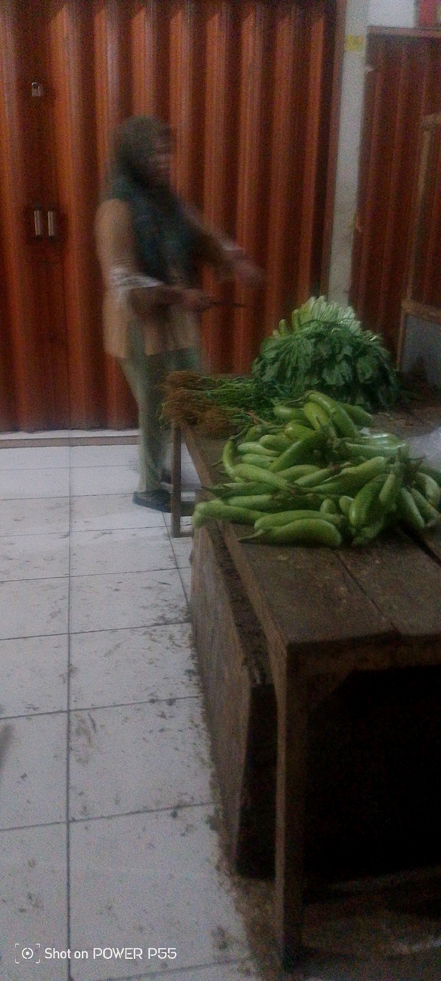 An indoor market stall is visible, with a person in the background blurred and out of focus. They are wearing a light-colored shirt and dark pants and a headscarf. A wooden table is in the foreground, laden with a pile of green vegetables, possibly cucumbers or eggplants, and bundles of leafy greens. The floor is tiled. The stall appears to be behind a large, corrugated metal shutter door. The lighting is dim, and the image quality is low. The camera's watermark "Shot on POWER P55" is present in the bottom left corner.