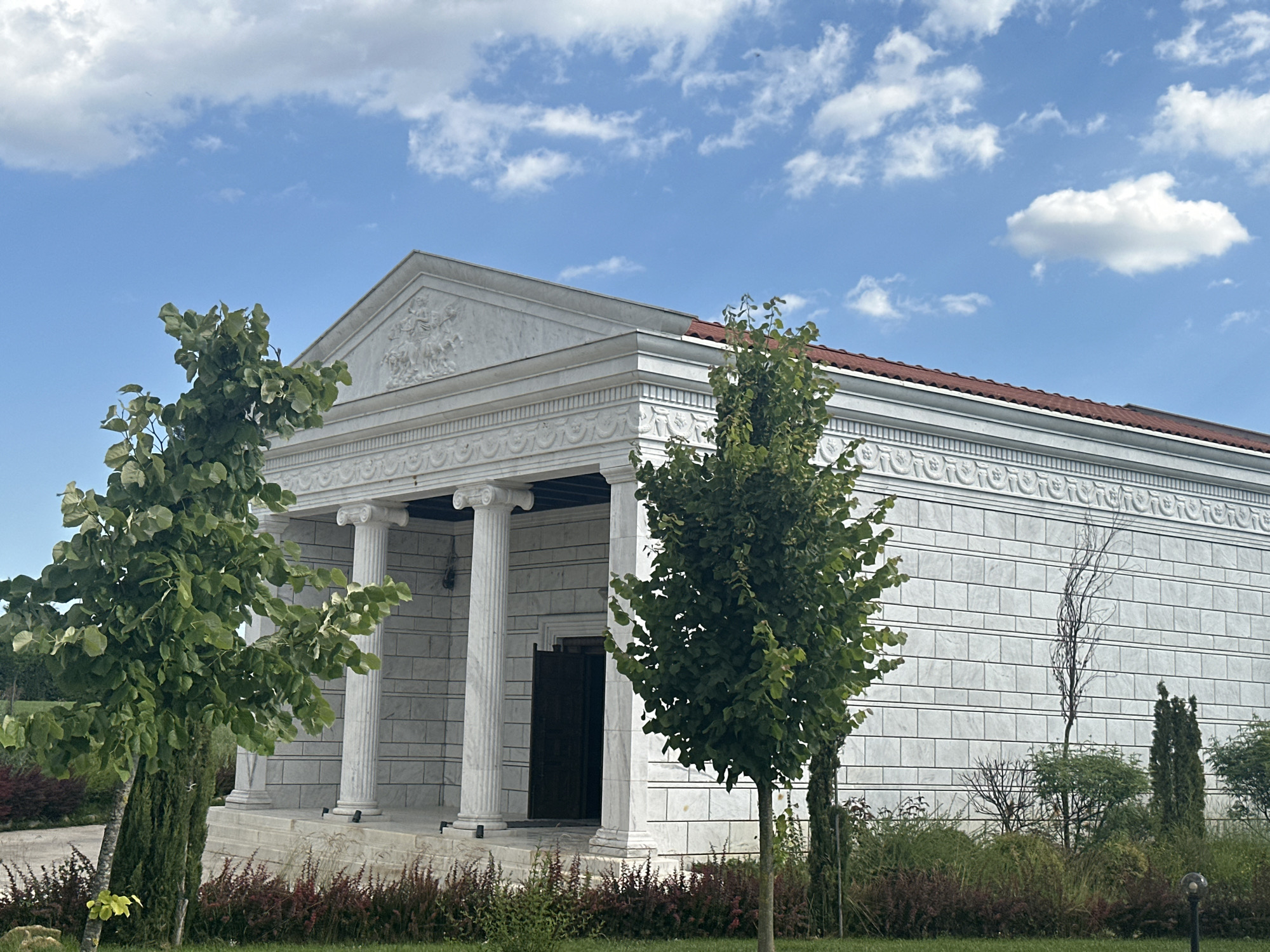 The image captures a facade of a white building with classical architectural elements. The building has a triangular pediment adorned with a relief sculpture, and is supported by white columns. The walls are made of large, light-colored rectangular blocks, possibly marble. A dark wooden door is visible between the columns. In front of the building, there are two leafy green trees, one on the left and one on the right, with a sparse arrangement of other bushes and plants in the foreground. The sky above is blue with scattered white clouds. The environment appears to be an outdoor setting with manicured landscaping. The architectural style suggests a neoclassical or ancient Greek influence. This building is located in the city of Neofit Rilski, Bulgaria.