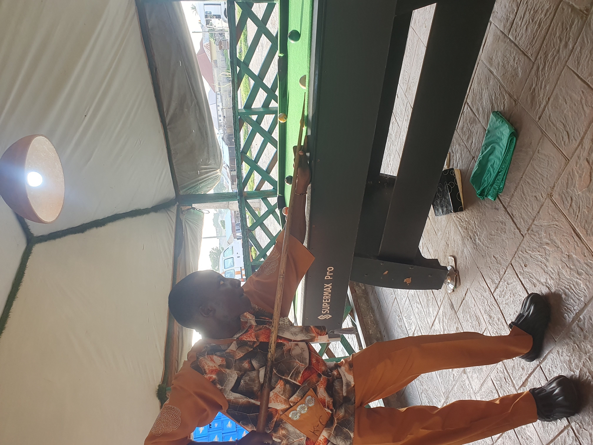 In Lokoja, Nigeria, a man is playing pool under a covered outdoor area with a thatched roof. He is wearing an orange patterned shirt and orange pants, and black clogs. He is holding a pool cue and is in the process of taking a shot. The pool table is green and has several balls on it, including a white cue ball, a red ball, a black ball, a yellow ball, and a green ball. The table has "SUPERMAX Pro" written on its side. In the background, there is a wooden fence and some buildings. The ground is paved with textured tiles. A green folded cloth and a black and white book are on the ground under the pool table.