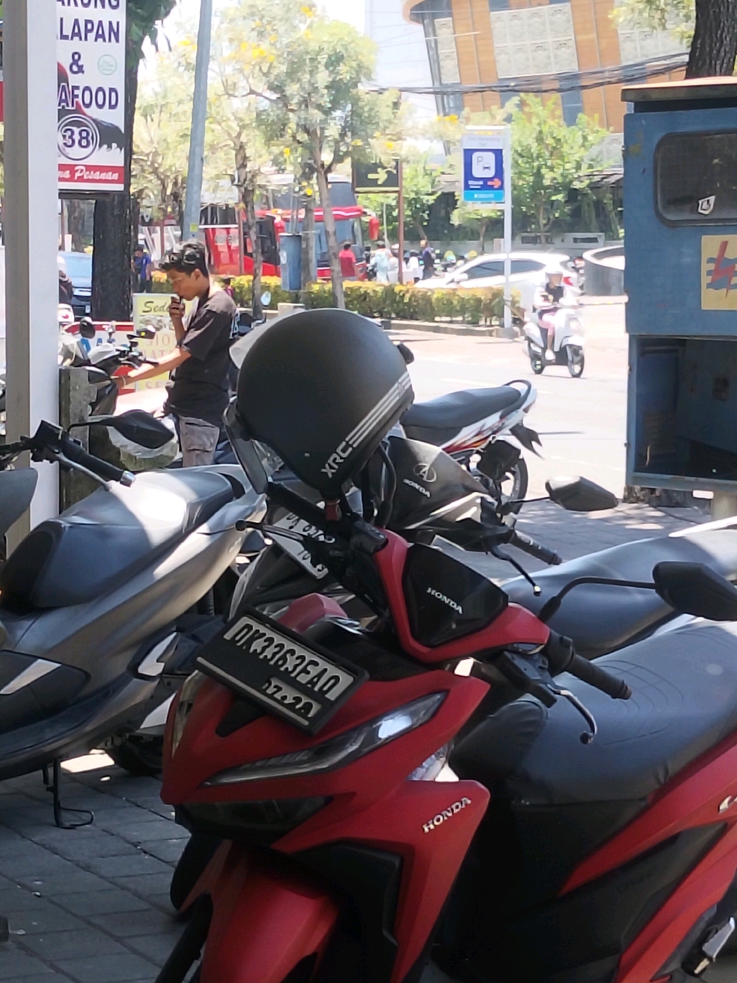 The image captures a street scene in Kuta, Indonesia, with a focus on a man holding a smartphone and standing next to motorcycles. The man, wearing a dark t-shirt and light shorts, appears to be engaged with his phone. Several motorcycles are parked, including a red one in the foreground with a license plate that reads "DK3363FAQ" and a date "07.20". A black helmet with the "XRC" logo sits atop the handlebars of the red motorcycle. In the background, a blue parking sign with a "P" symbol and an arrow is visible, along with trees, buildings, and other vehicles. The overall environment suggests a busy urban setting, likely a commercial area or parking lot.