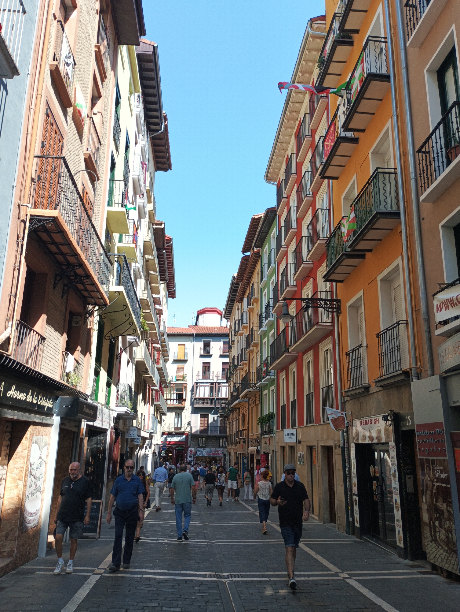 This is a street-level, eye-level, outdoor, full-shot photograph taken in Pamplona, Spain, on a sunny day, looking down a cobblestone street lined with colorful buildings. The buildings have multiple stories, balconies with wrought-iron railings, and open windows. Several flags, primarily red, white, and green, are visible hanging from balconies. People are walking along the street, some in groups and some individually. Shops and businesses with awnings and signage are present on the ground floor of the buildings. The sky is clear and blue. The street appears to be a pedestrian area.