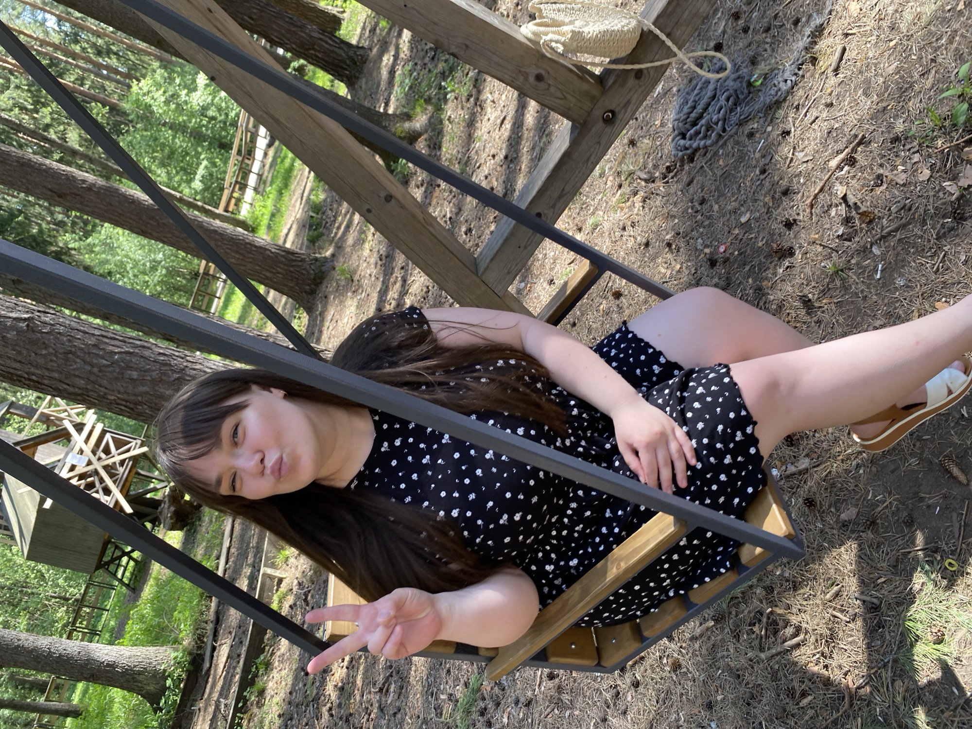 A woman is sitting on a swing set outdoors. She is wearing a black dress with a white floral pattern and sandals. She is holding up two fingers in a peace sign and puckering her lips. The location appears to be a wooded area, possibly a park or recreational area near Votkinsk, Russia, due to the presence of pine trees. The swing set is made of wood and metal. A small straw purse is visible hanging on the wooden structure behind the swing. The ground is covered with pine needles, twigs, and other natural debris. A wooden platform structure is also visible in the background.