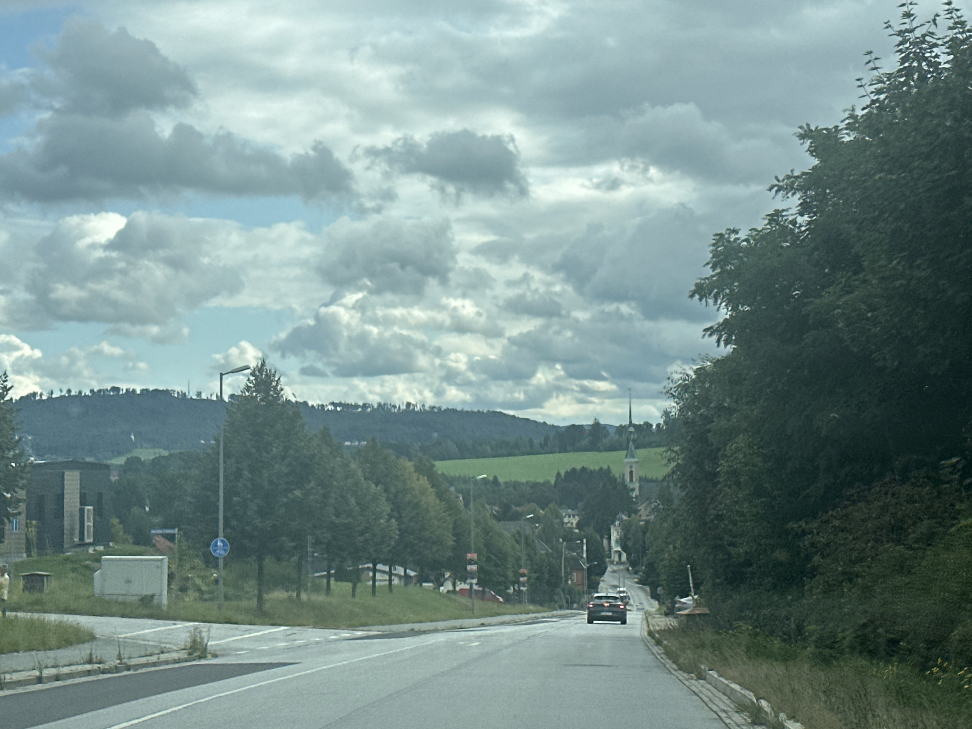 The image captures a scenic view of a rural road in Großpostwitz, Germany, on a cloudy day. The road curves gently ahead, with a dark-colored car in the distance. On the right side of the road, dense green trees line the landscape, partially obscuring a steep embankment. To the left of the road, there is a grassy verge and a few buildings. Further in the distance, rolling green hills are visible under a dramatic sky filled with white and grey clouds. A church steeple with a green roof pierces the skyline, indicating the presence of a village or town nestled in the valley. The overall atmosphere is one of a quiet, picturesque countryside.